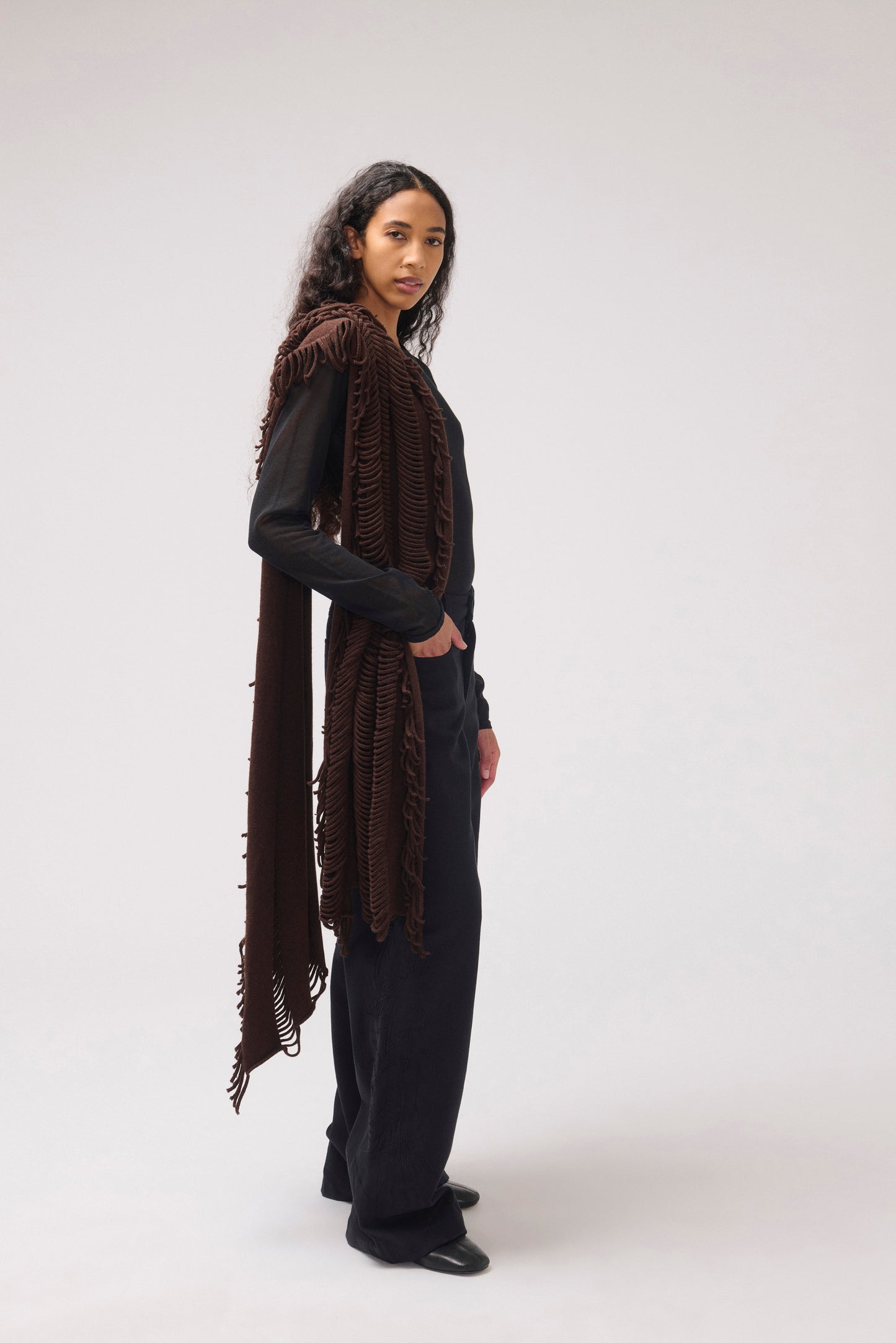 A model stands in profile wearing a long, dark brown fringed  shawl over a black outfit, photographed against a plain light gray studio background.