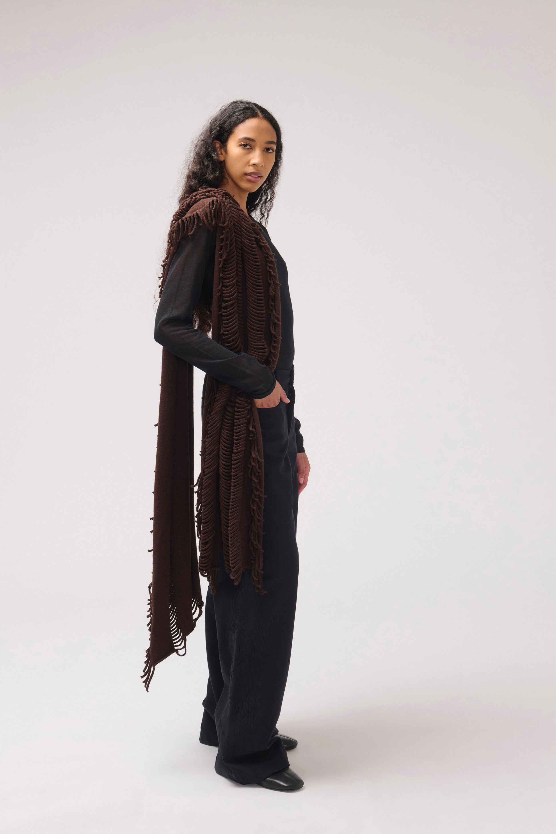 A model stands in profile wearing a long, dark brown fringed  shawl over a black outfit, photographed against a plain light gray studio background.