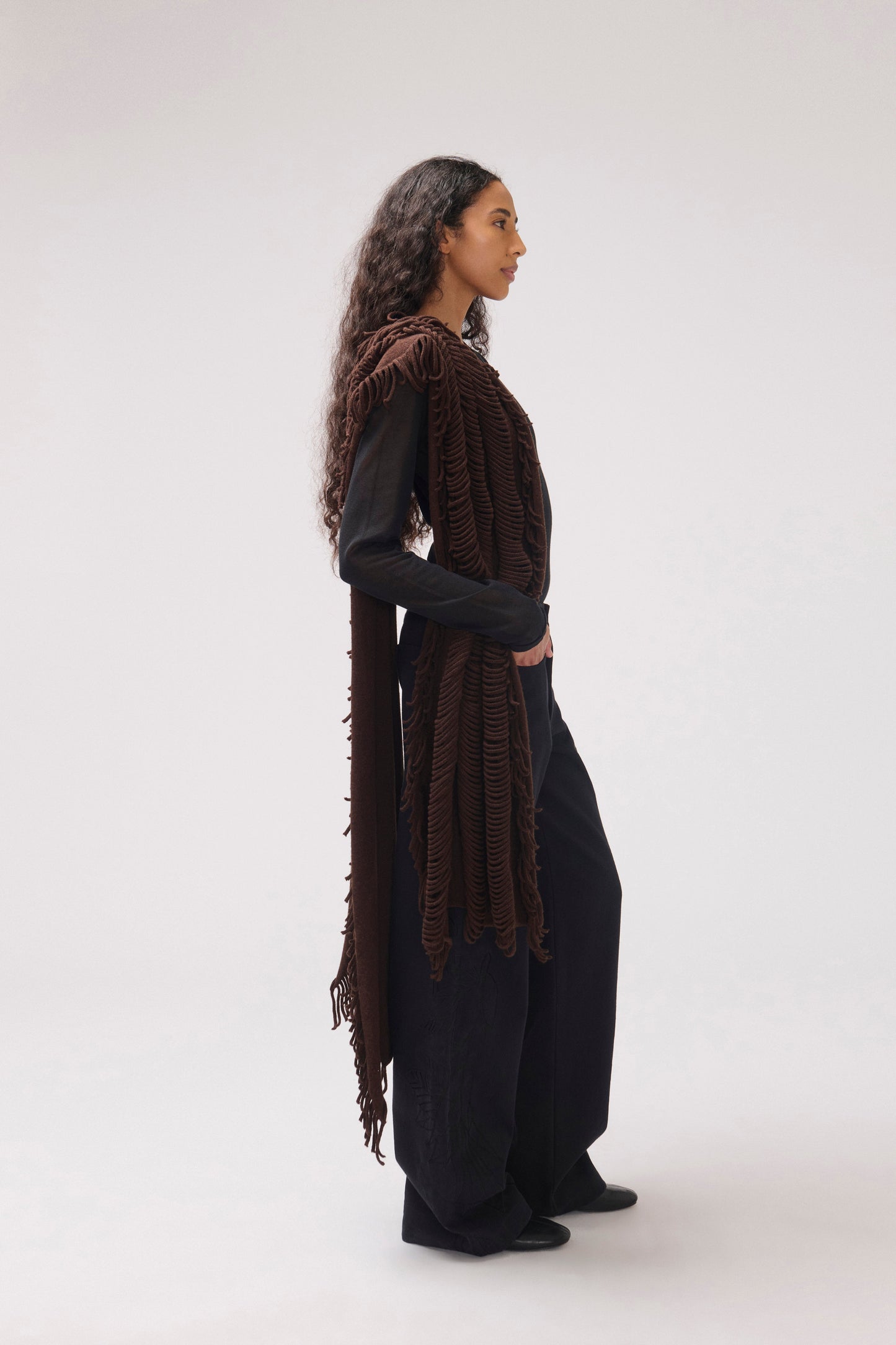 A model is shown in profile wearing a dark brown fringed scarf over a black long-sleeve top and black wide-leg pants, photographed against a seamless light gray studio background.