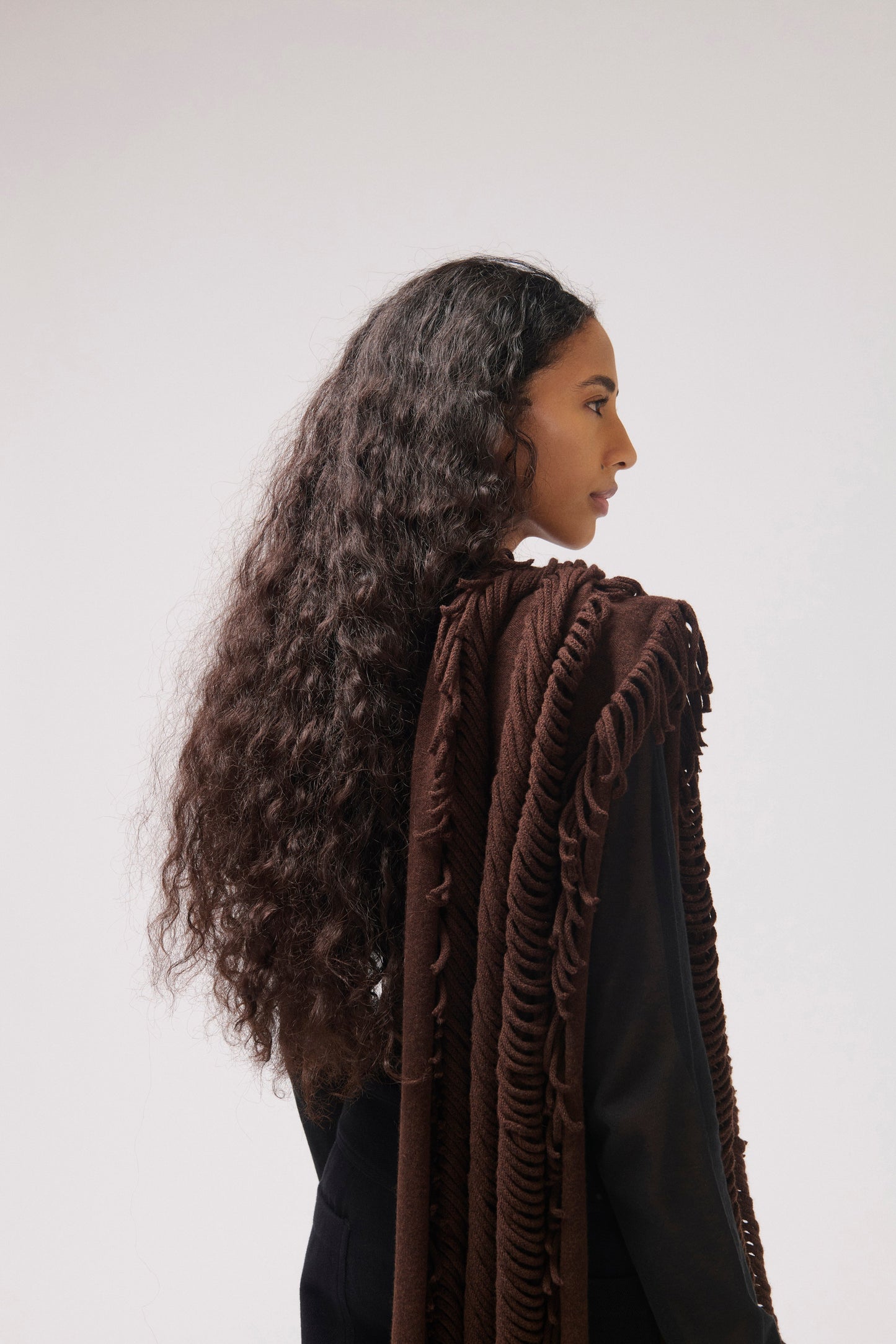 A model with long hair is shown in profile against a light gray studio background. She is wearing a dark brown fringed shawl draped over her shoulders and a black top.