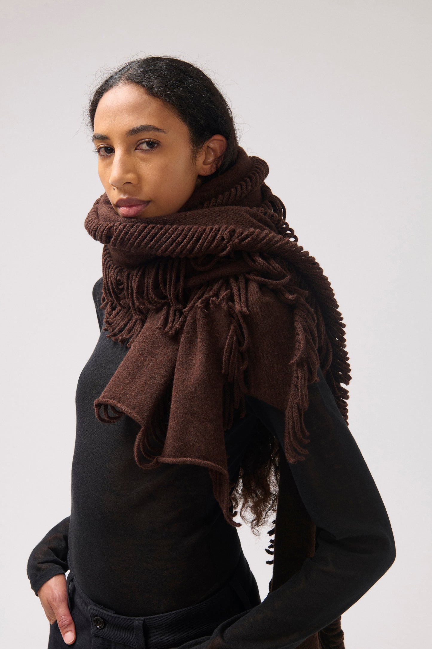 Model facing the camera wearing a black top and brown wool scarf with fringes.