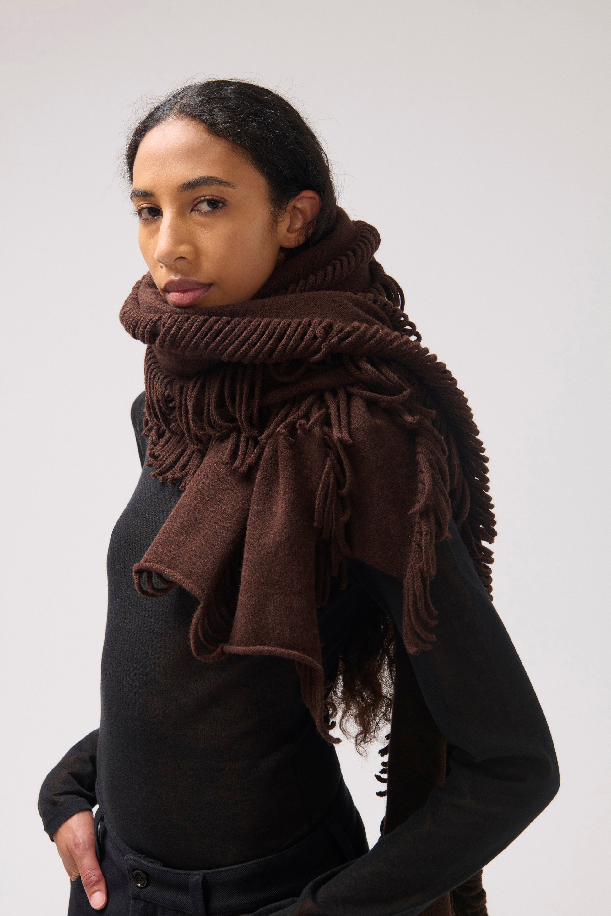 Model facing the camera wearing a black top and brown wool scarf with fringes.