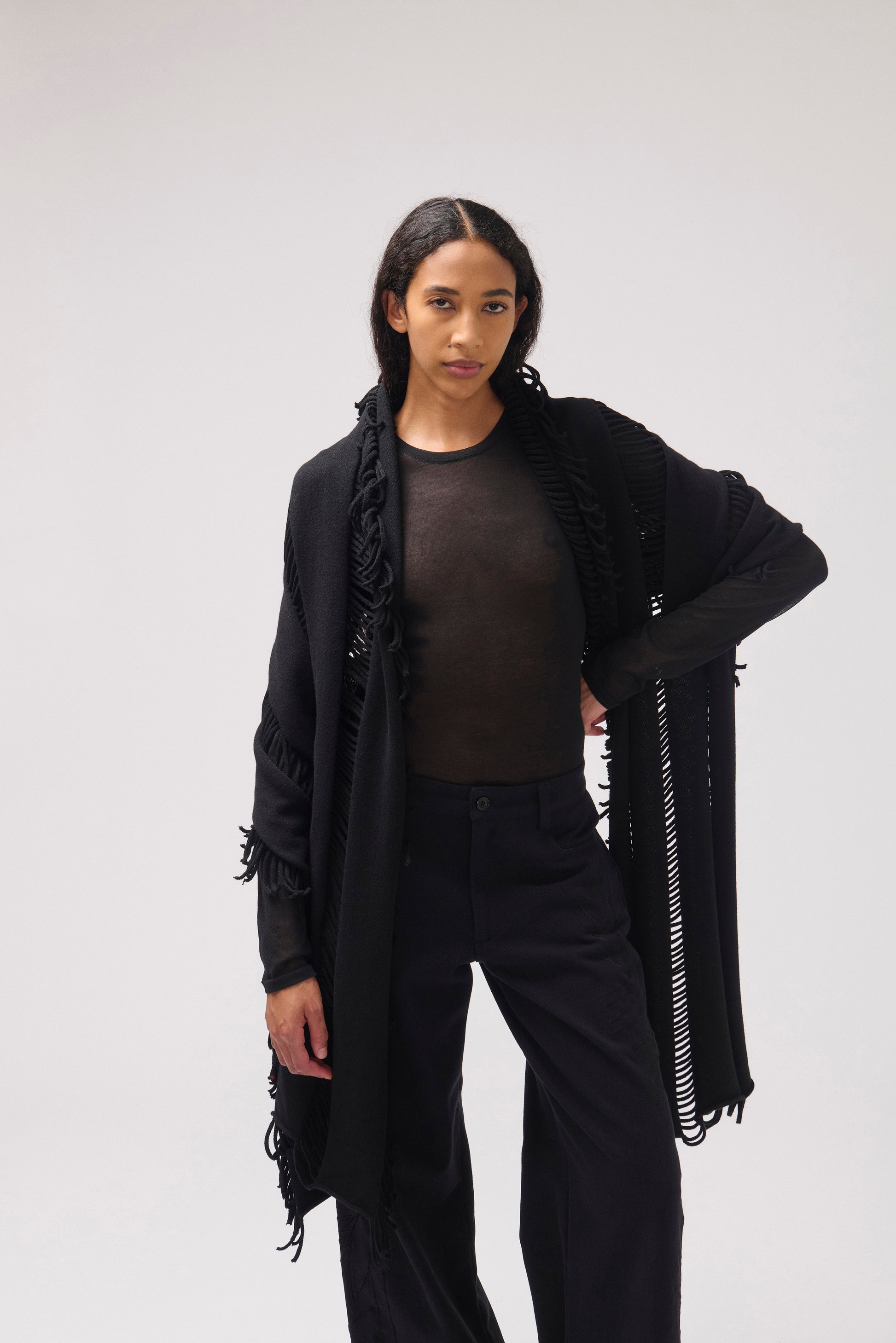 A model wearing a black fringed shawl over a black top and black pants, photographed against a neutral light gray studio background. Her arm is placed on her waist.