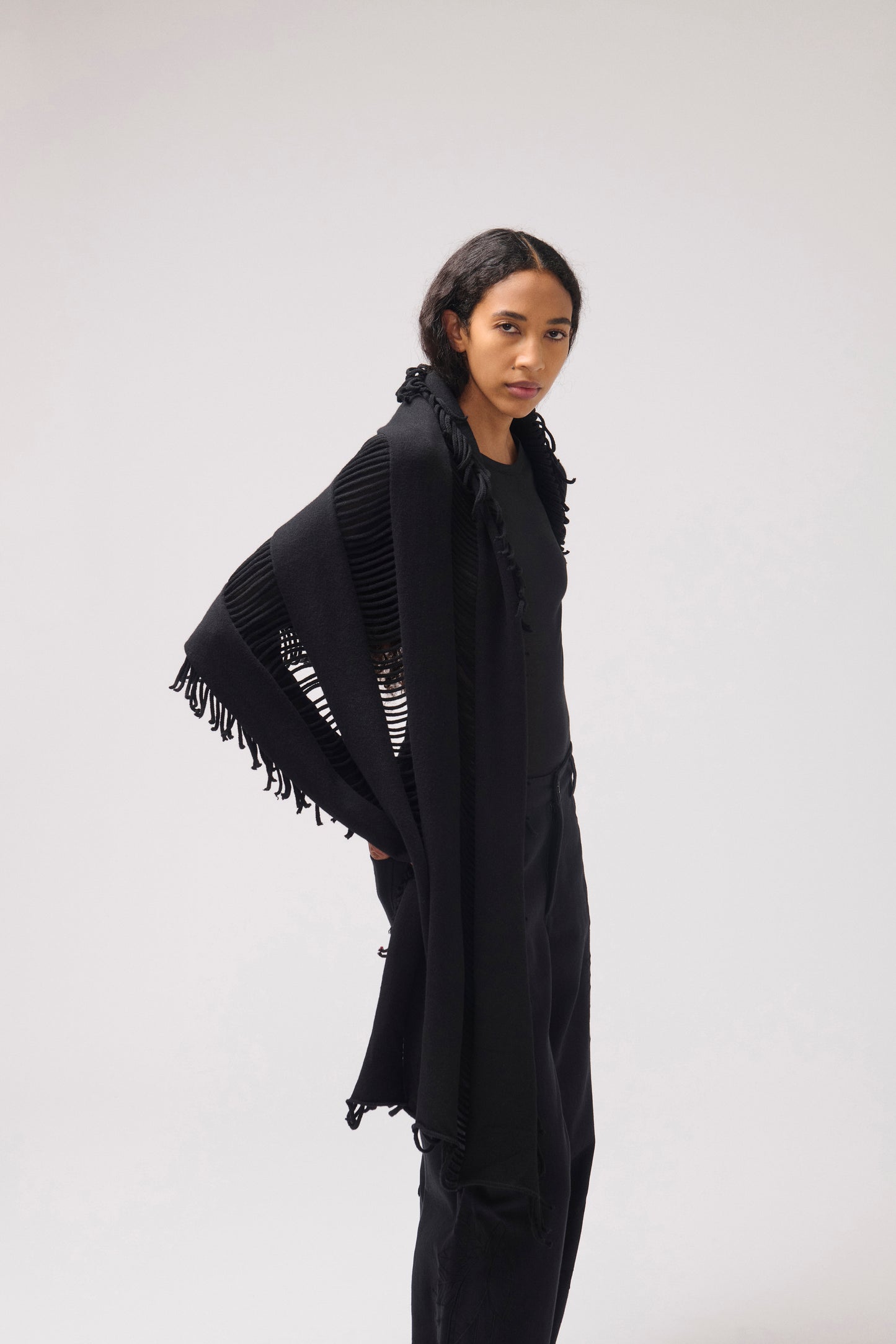 A model wearing a black fringed shawl over a black top and black pants, photographed against a neutral light gray studio background.