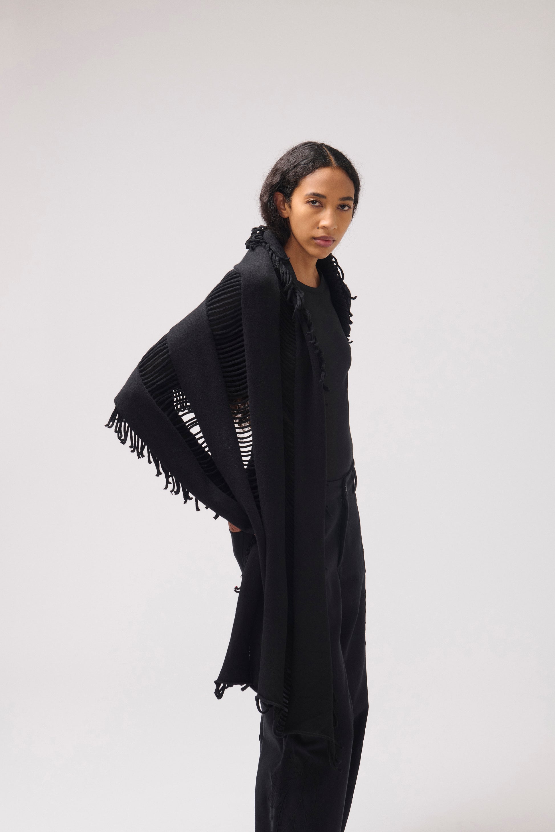 A model wearing a black fringed shawl over a black top and black pants, photographed against a neutral light gray studio background.