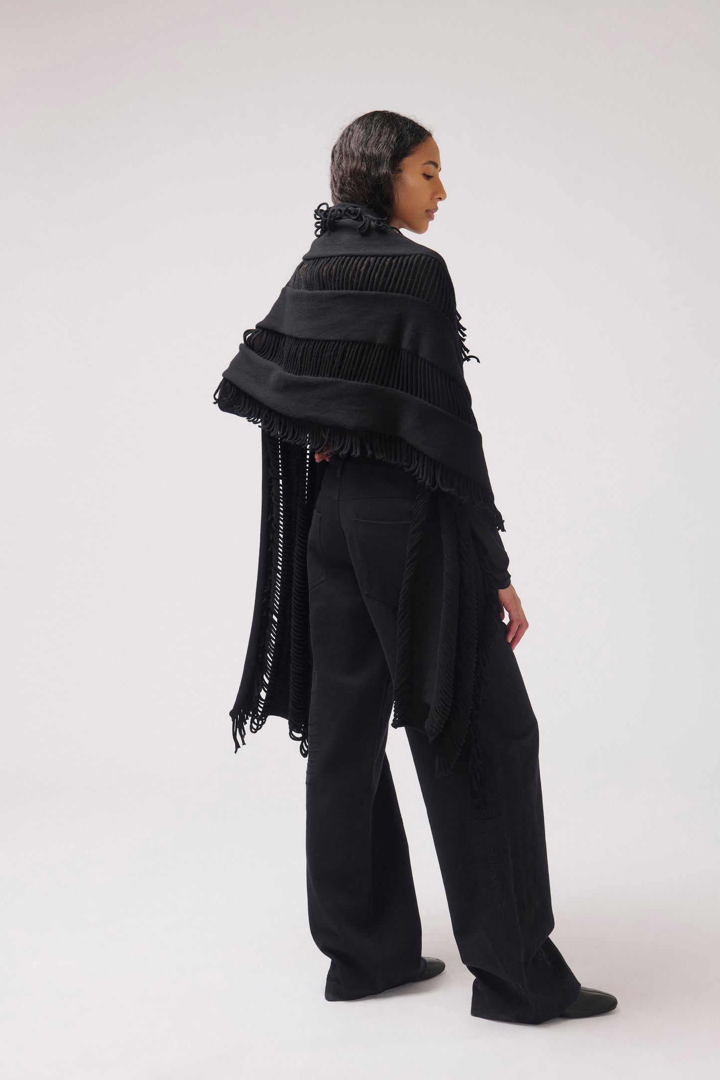 A model shown from the back wearing a black fringed shawl draped over wide-leg black trousers, photographed against a seamless light gray studio background.