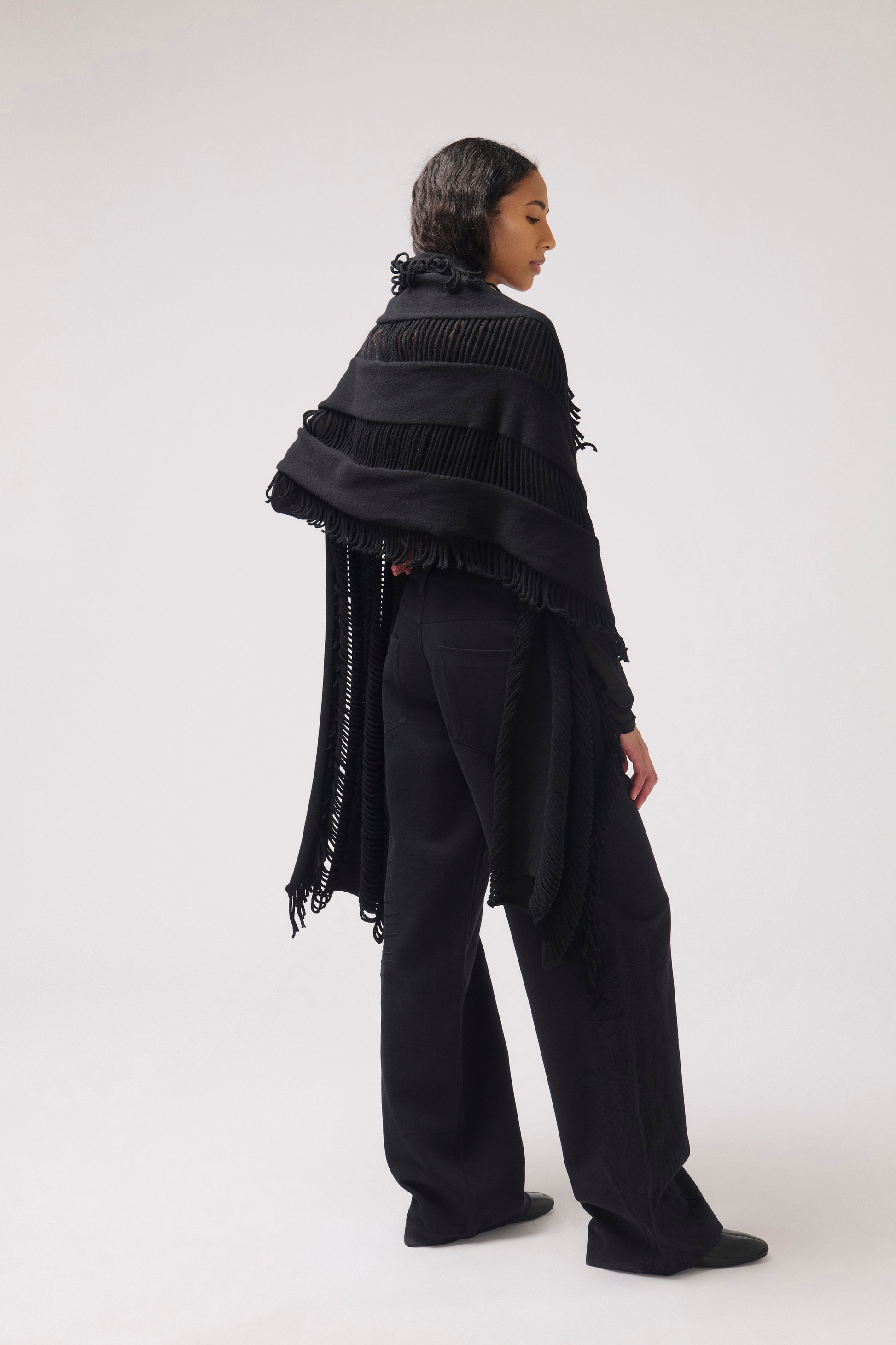 A model shown from the back wearing a black fringed shawl draped over wide-leg black trousers, photographed against a seamless light gray studio background.