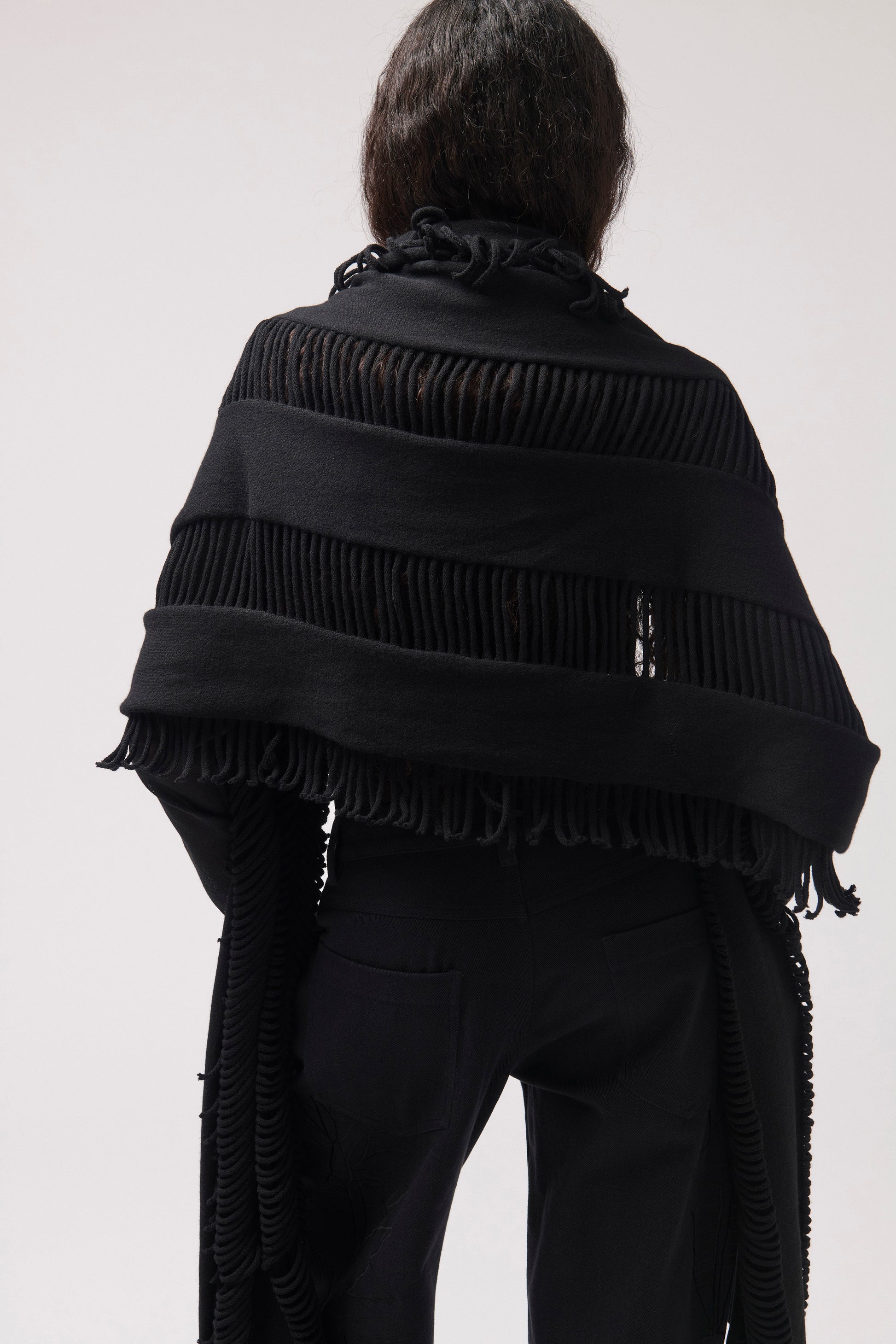 A person shown from the back wearing a black fringed shawl  draped over the shoulders, paired with black pants, against a plain light gray studio background.