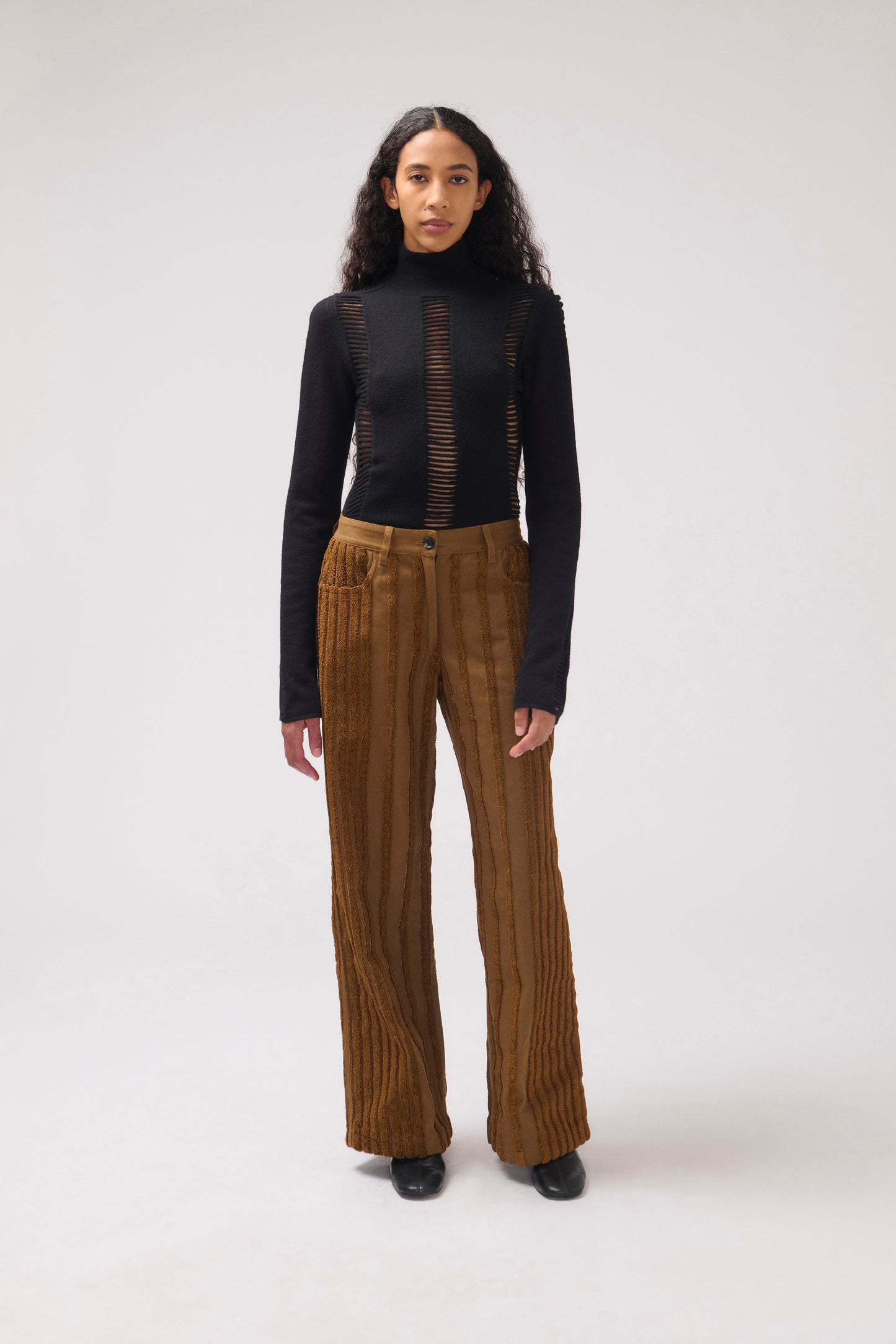 A model stands against a light gray studio background wearing a black long-sleeve turtleneck top with vertical brown mesh panels and high-waisted brown pleated wide-leg trousers.