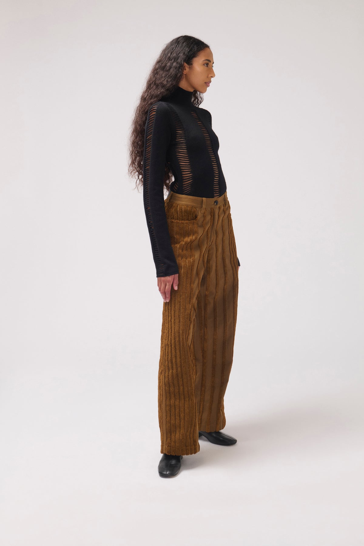 A model is shown from a side profile wearing a black long-sleeve knit top with sheer vertical panels and high-waisted, wide-leg brown corduroy trousers, standing against a plain light gray studio background.