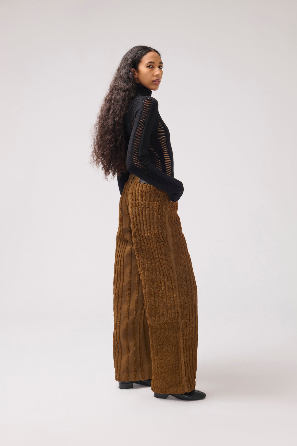 A model is shown from a side-back angle wearing a black long-sleeve top with sheer mesh panels and wide-leg brown corduroy trousers, standing against a plain light gray studio background.