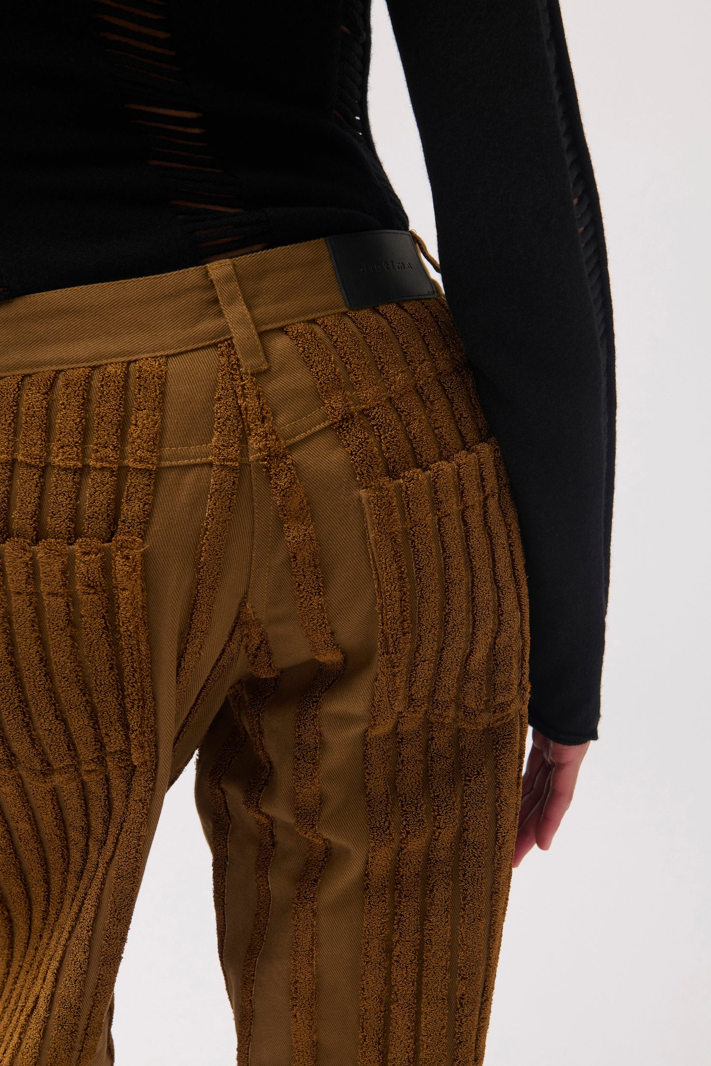 Close-up of a person wearing textured brown ribbed trousers with a black waistband and black long-sleeve top against a light gray studio background.