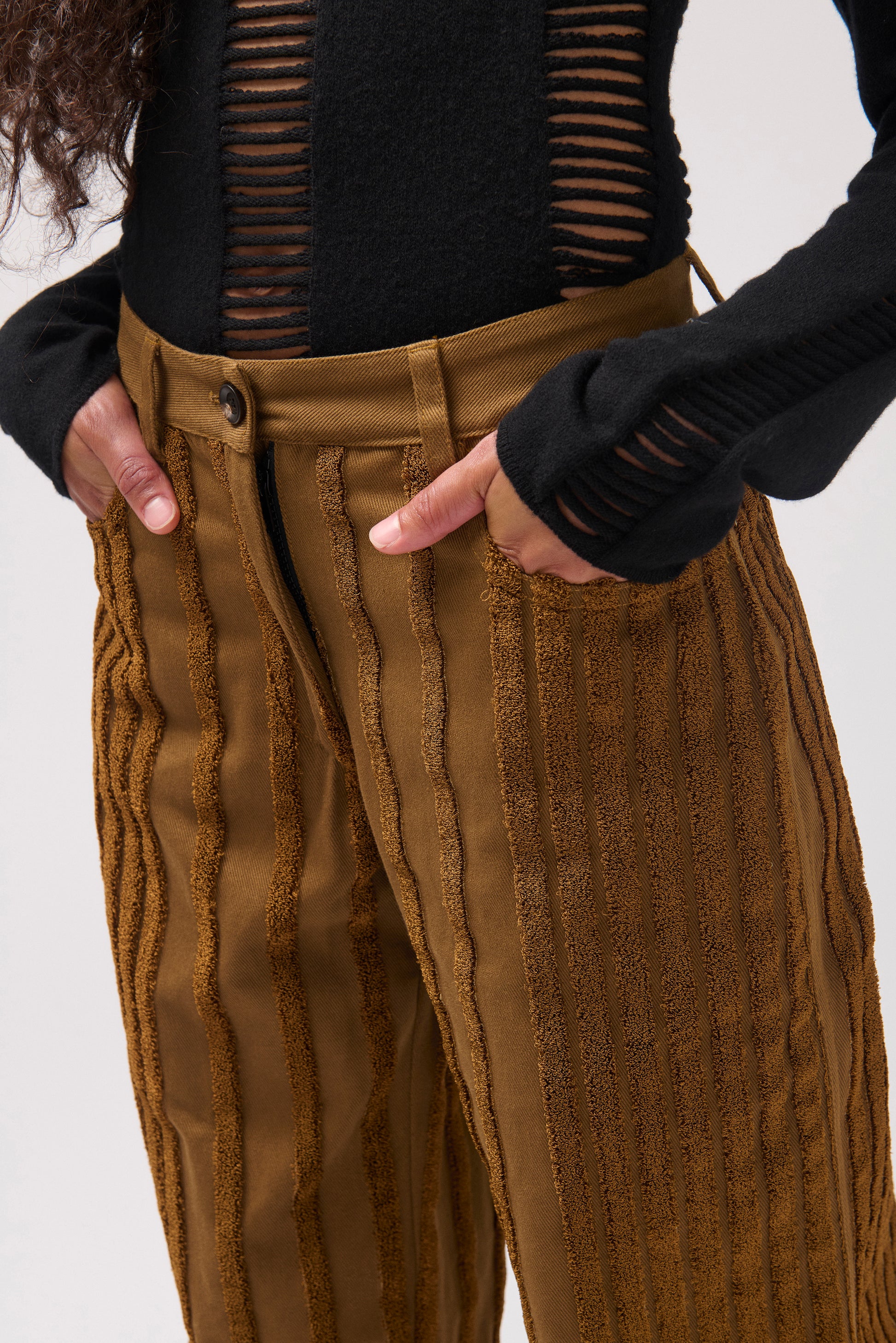 Close-up of a person wearing brown textured knit pants with vertical ribbed stripes and a black ribbed top with horizontal cutout details, shown against a light gray studio background.