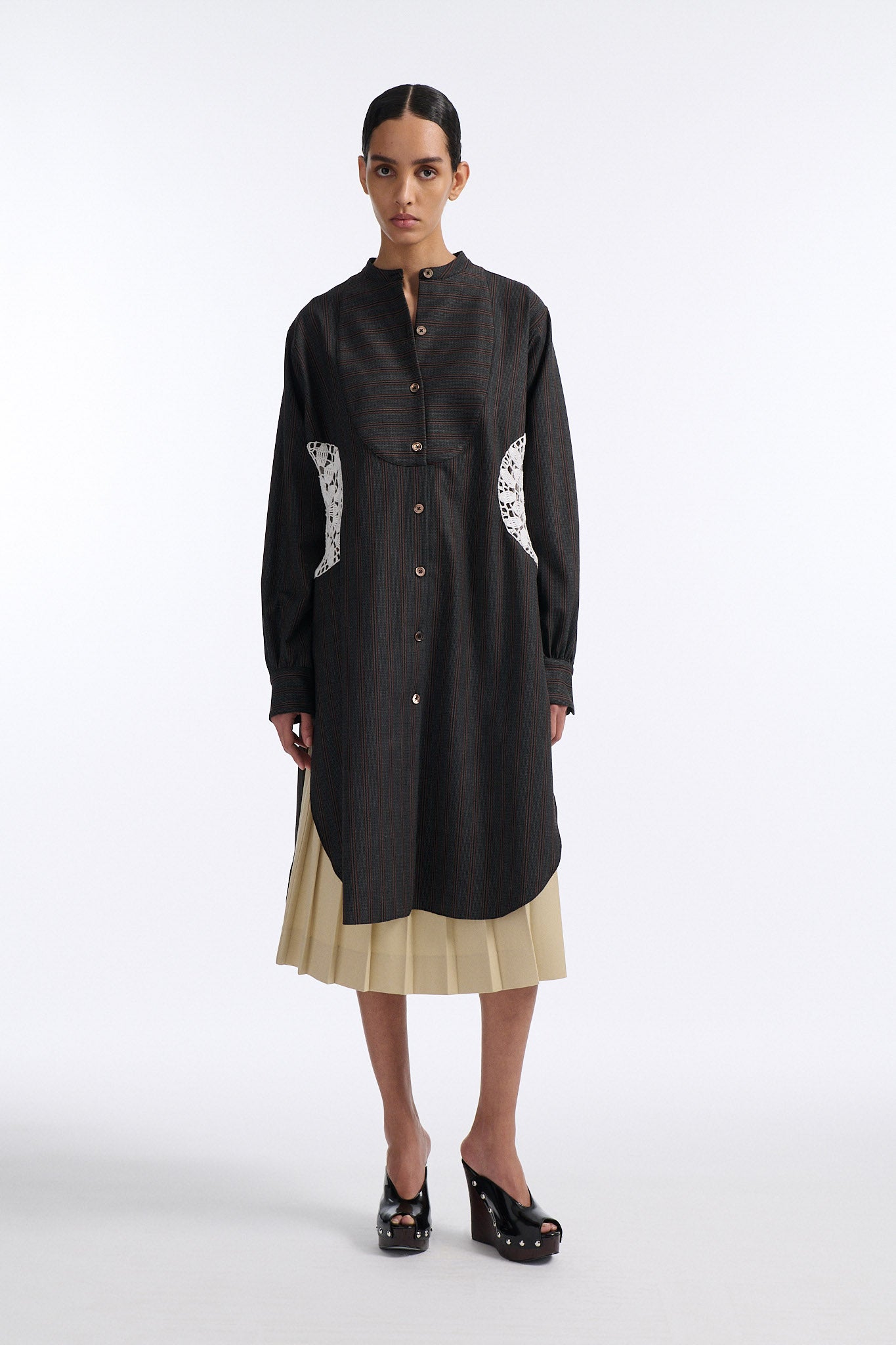 Female model with slicked back black hair in a bun wears charcoal grey button down shirt dress with white circle crochet insert. Shirt has thin burgundy vertical stripes. Model also wears beige pleated skirt and black wedges. 