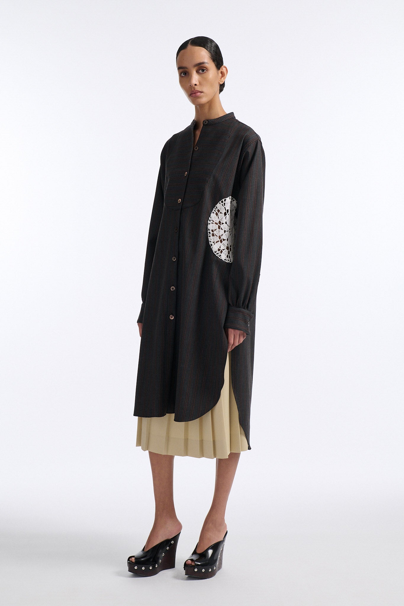 Female model with slicked back black hair in a bun wears charcoal grey button down shirt dress with white circle crochet insert. Shirt has thin burgundy vertical stripes. Model also wears beige pleated skirt and black wedges. 