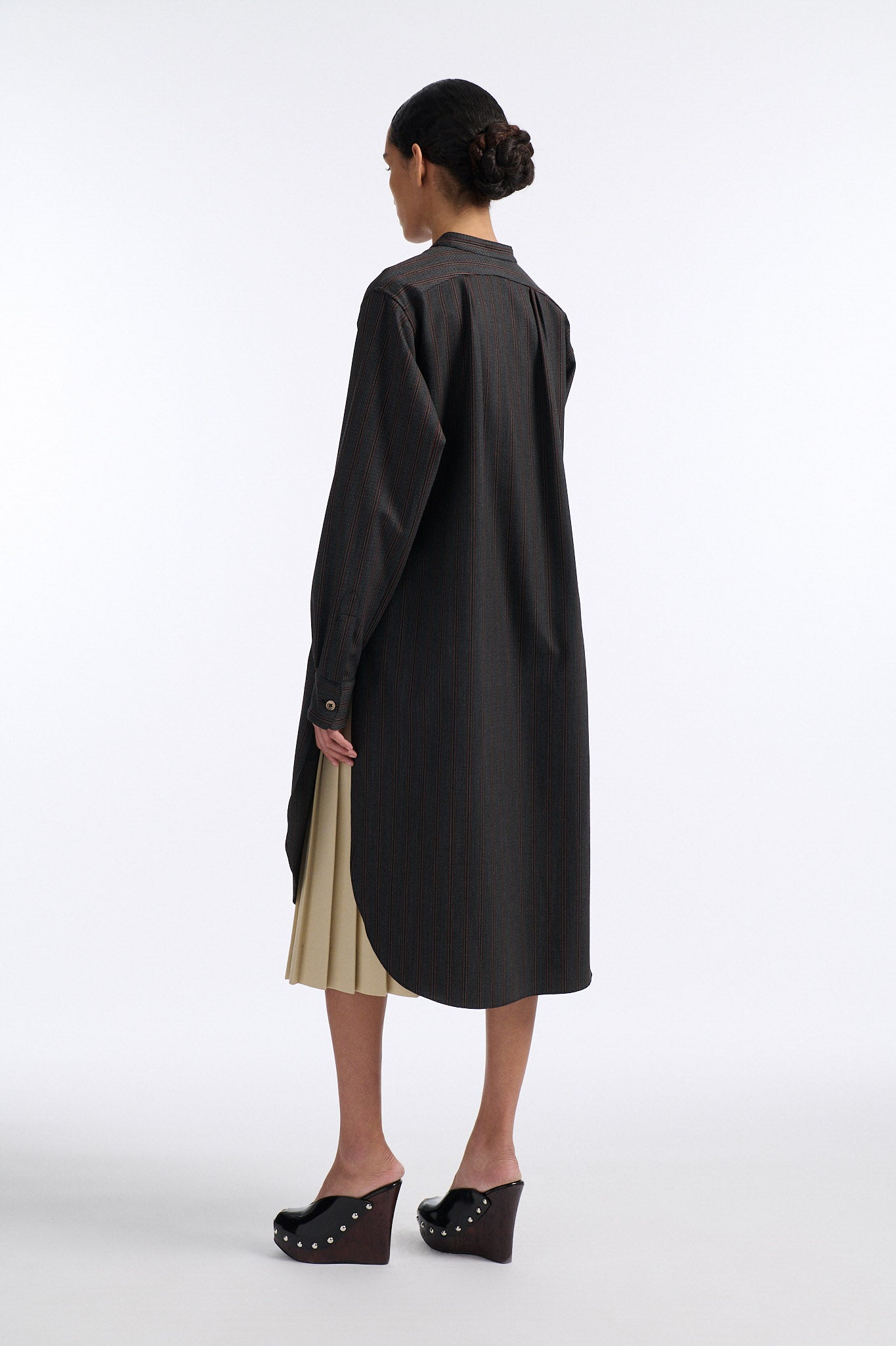 Female model with slicked back black hair in a bun wears charcoal grey button down shirt dress with white circle crochet insert. Shirt has thin burgundy vertical stripes. Model also wears beige pleated skirt and black wedges. 