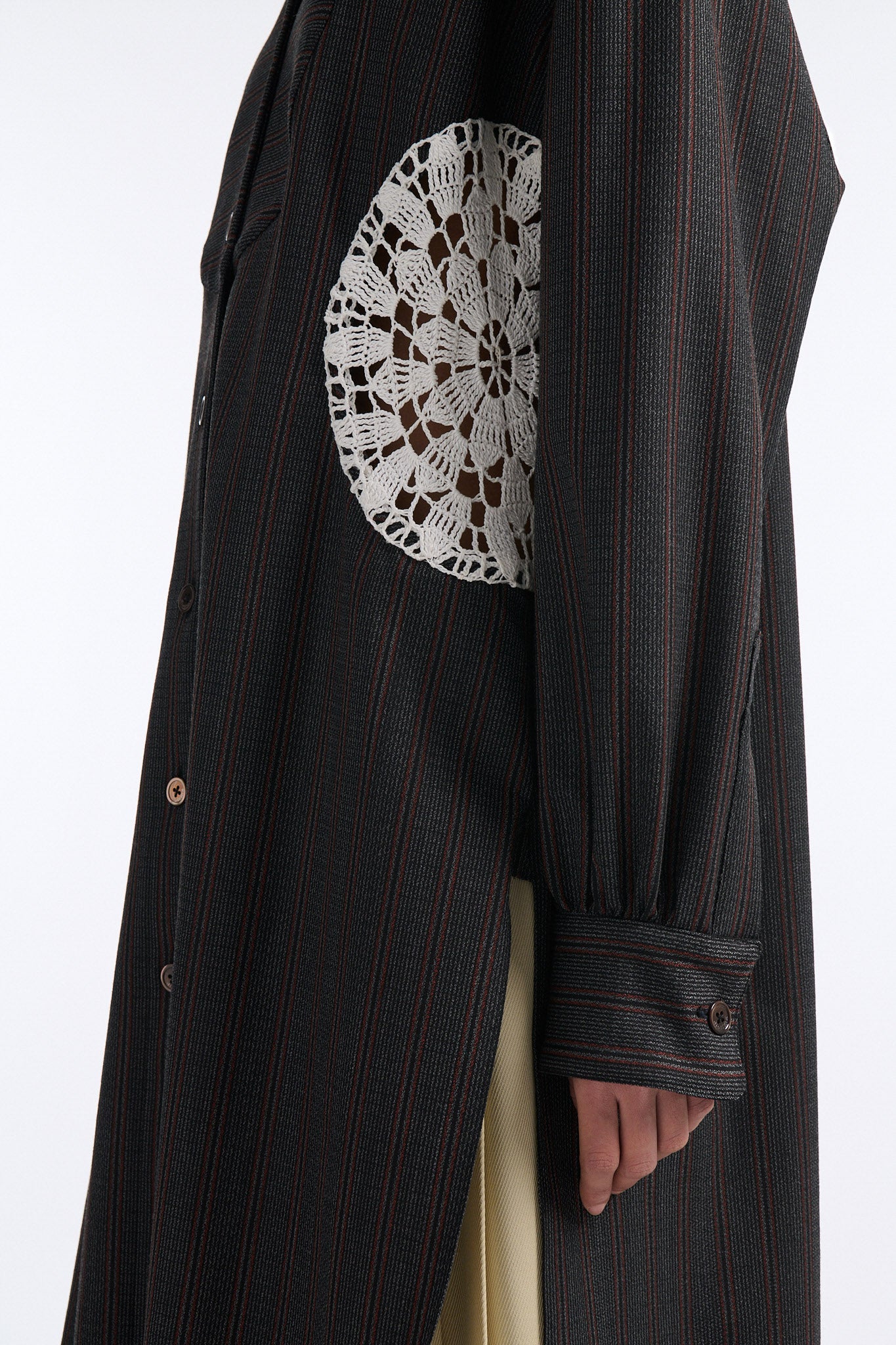Female model with slicked back black hair in a bun wears charcoal grey button down shirt dress with white circle crochet insert. Shirt has thin burgundy vertical stripes. Model also wears beige pleated skirt and black wedges. 