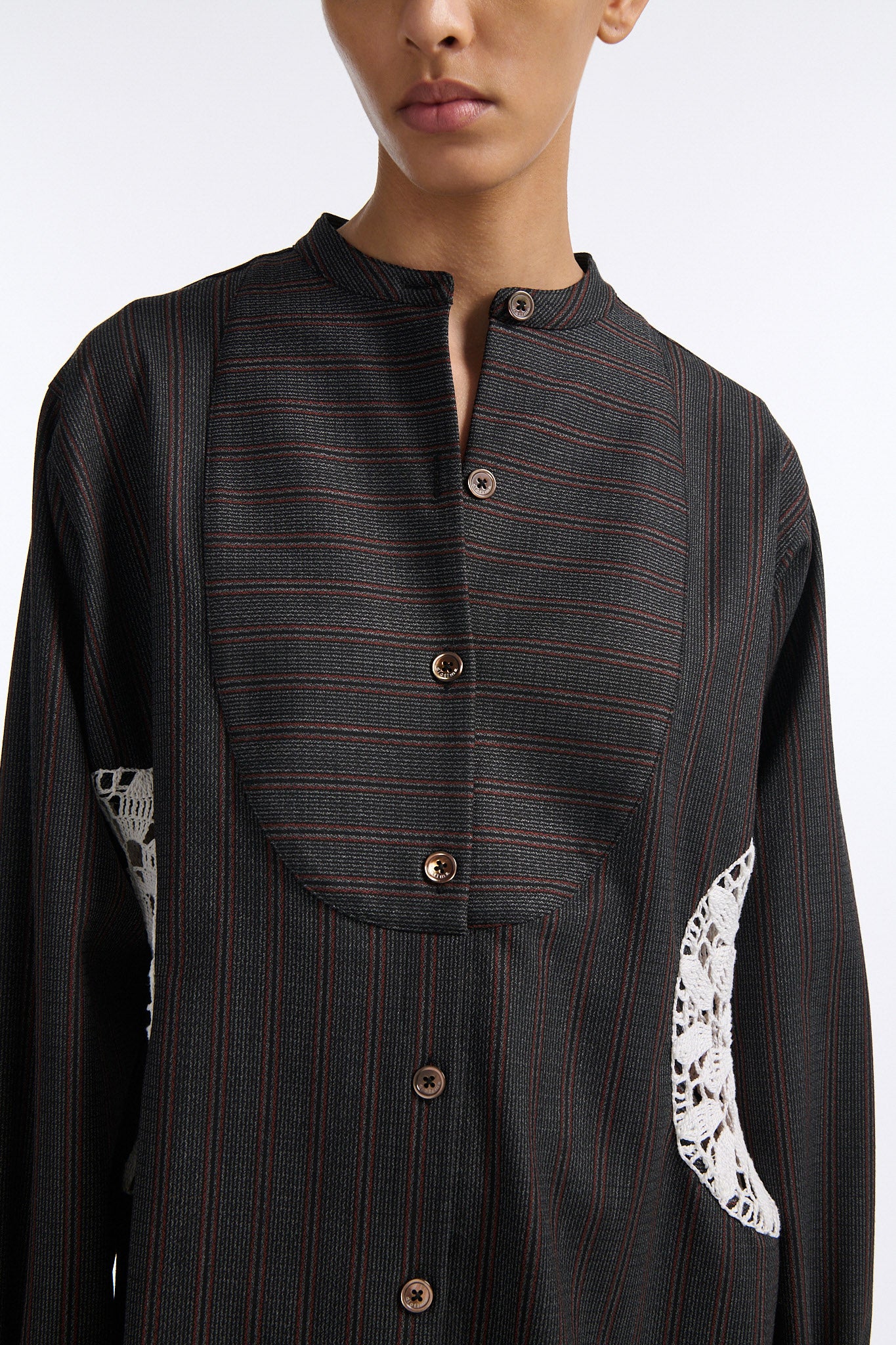 Female model with slicked back black hair in a bun wears charcoal grey button down shirt dress with white circle crochet insert. Shirt has thin burgundy vertical stripes. Model also wears beige pleated skirt and black wedges. 