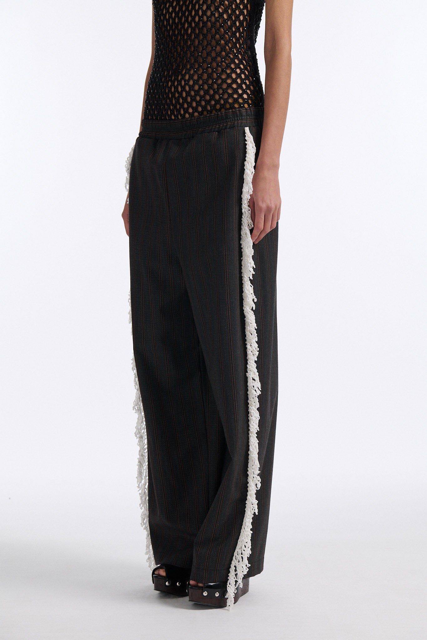 Female model with slicked back black hair in a bun wears back fine crystal embellished mesh tank with charcoal grey and burgundy vertical striped trousers. Trousers have elastic waist with white crochet fringe along either leg. 