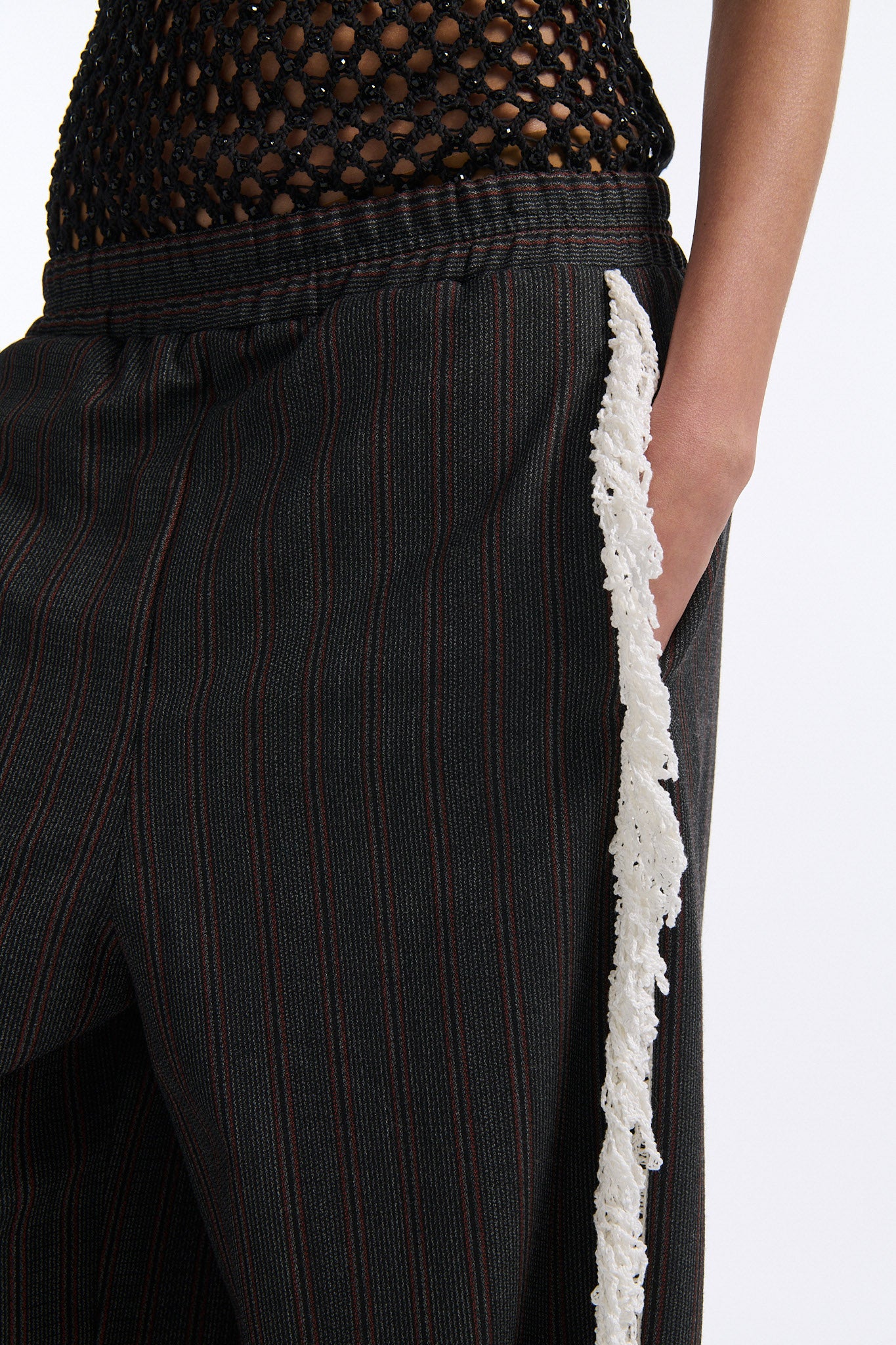 Female model with slicked back black hair in a bun wears back fine crystal embellished mesh tank with charcoal grey and burgundy vertical striped trousers. Trousers have elastic waist with white crochet fringe along either leg. 