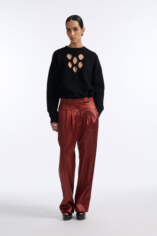 Female model with slicked back black hair in a bun wears black crewneck long sleeve sweater with 5 hole cutout at center of chest. Model also wears shiny rust red pleated trousers with black wedges.