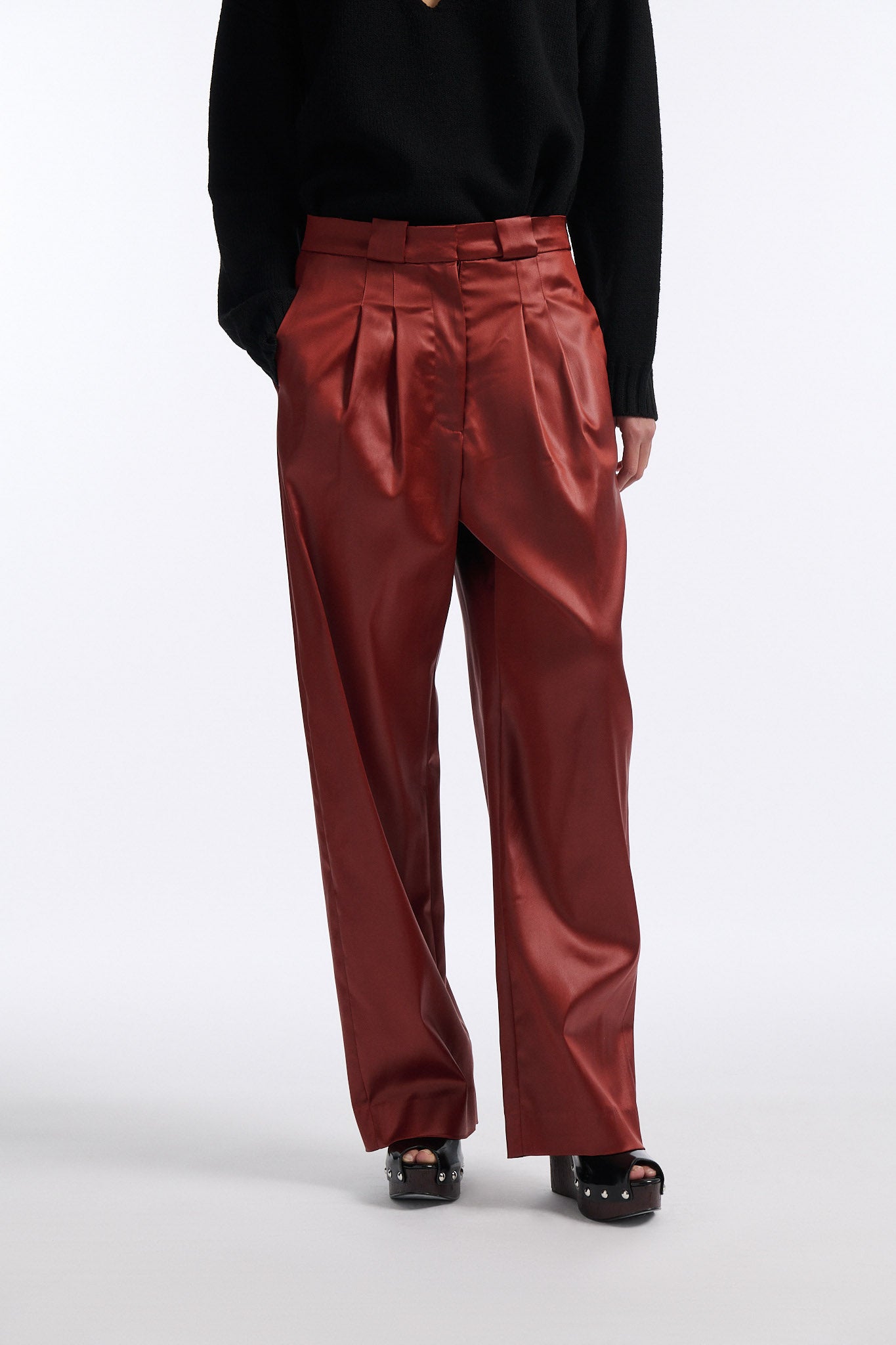 Female model with slicked back black hair in a bun wears black crewneck long sleeve sweater with 5 hole cutout at center of chest. Model also wears shiny rust red pleated trousers with black wedges.
