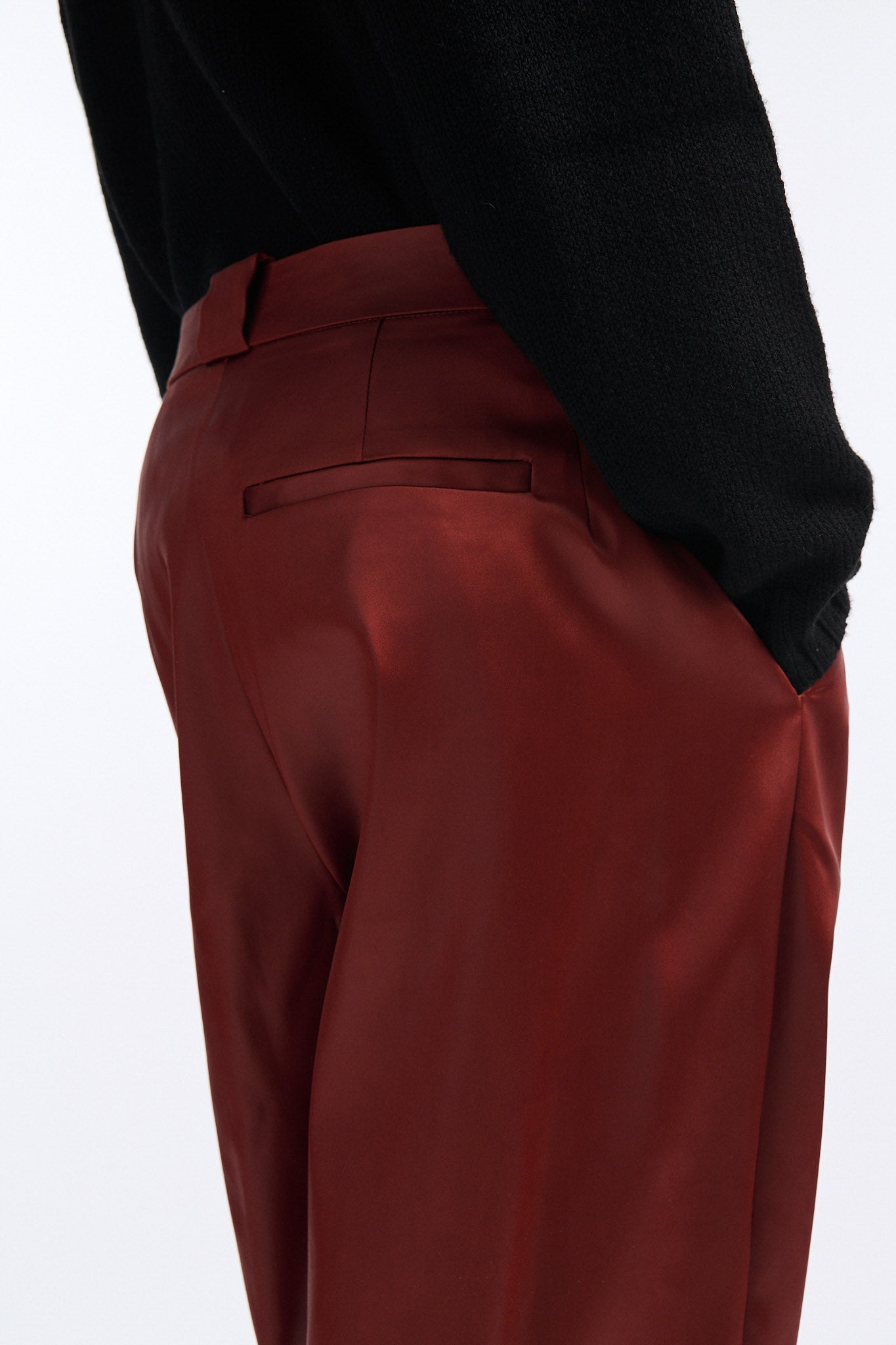Female model with slicked back black hair in a bun wears black crewneck long sleeve sweater with 5 hole cutout at center of chest. Model also wears shiny rust red pleated trousers with black wedges.