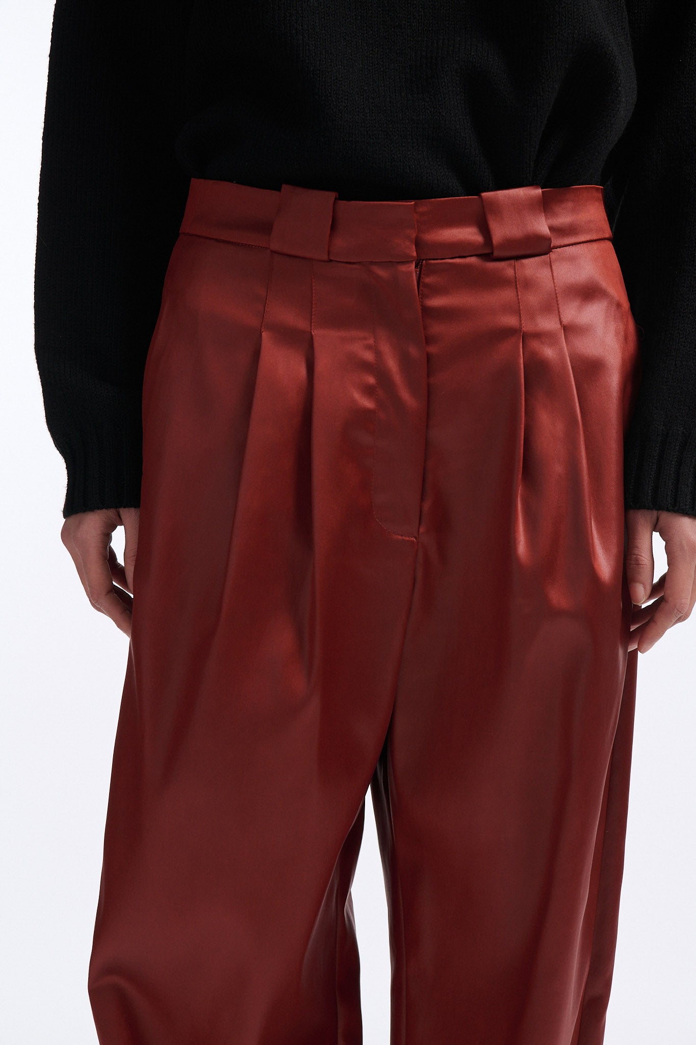 Female model with slicked back black hair in a bun wears black crewneck long sleeve sweater with 5 hole cutout at center of chest. Model also wears shiny rust red pleated trousers with black wedges.