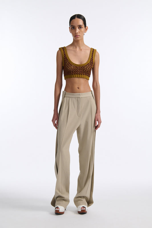 Female model with slicked back black hair in a bun wears cumin colored chunky mesh bralette with maroon hand-sewn crystals. Model also wears beige wide pleated trousers with additional panel at sides to increase volume. 