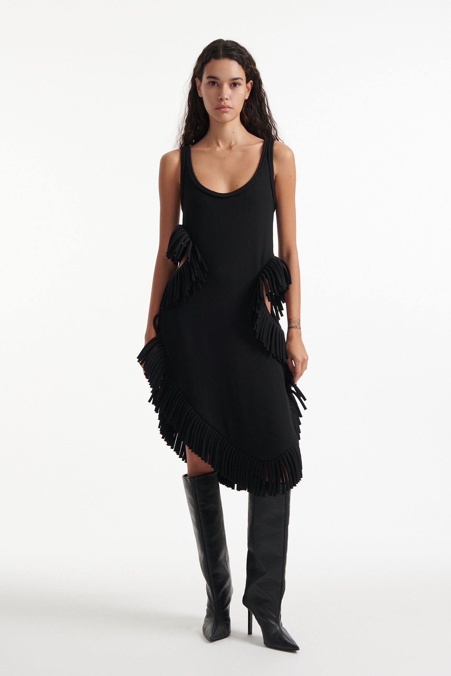 Female model wears black midi tank dress with large fringe openings at either side and scoop fringe hem with black high heel boots on white background.