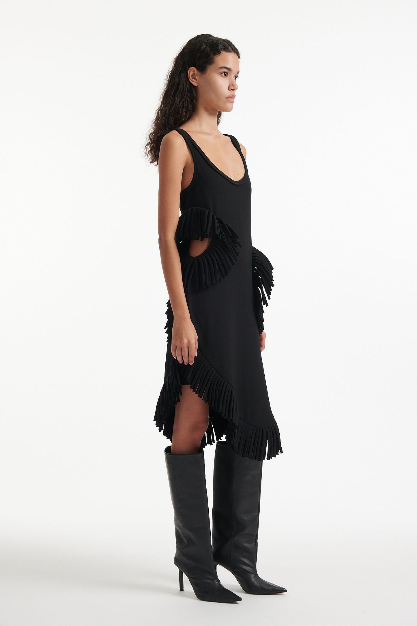 Female model wears black midi tank dress with large fringe openings at either side and scoop fringe hem with black high heel boots on white background.