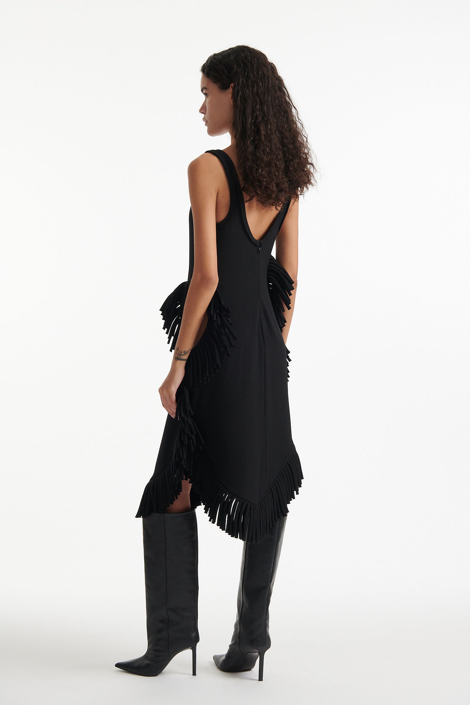 Female model wears black midi tank dress with large fringe openings at either side and scoop fringe hem with black high heel boots on white background.