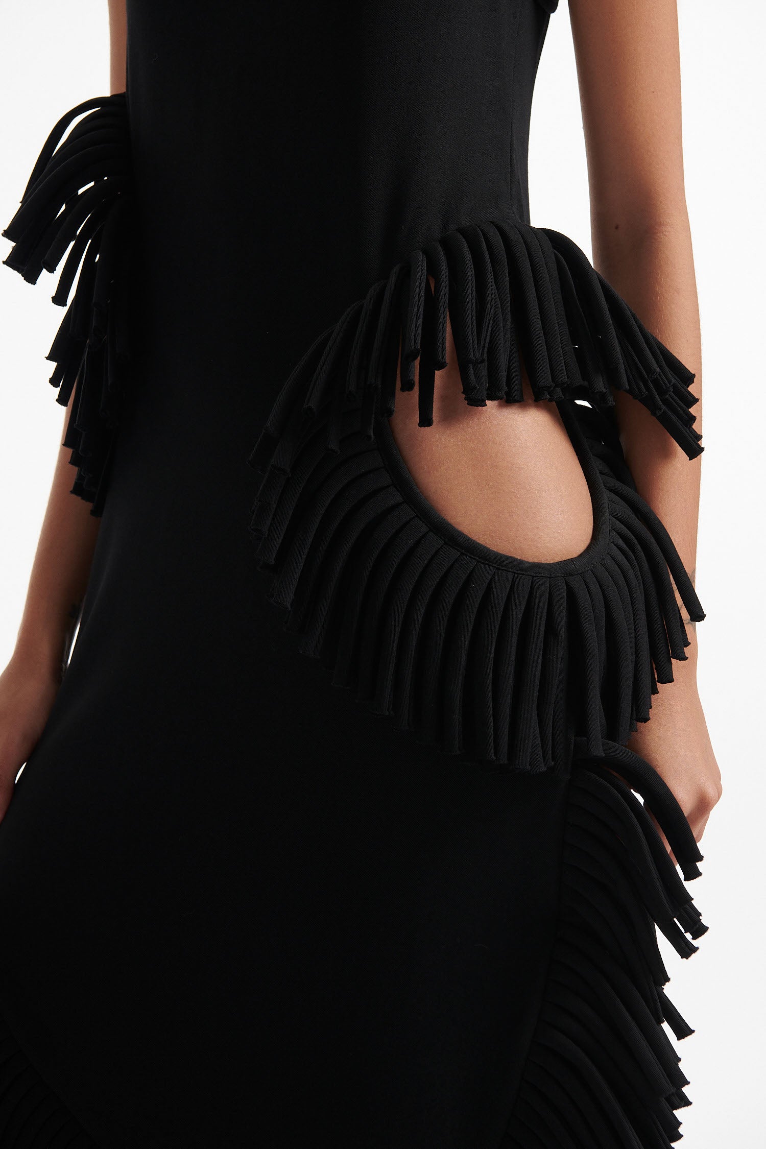 Female model wears black midi tank dress with large fringe openings at either side and scoop fringe hem with black high heel boots on white background.