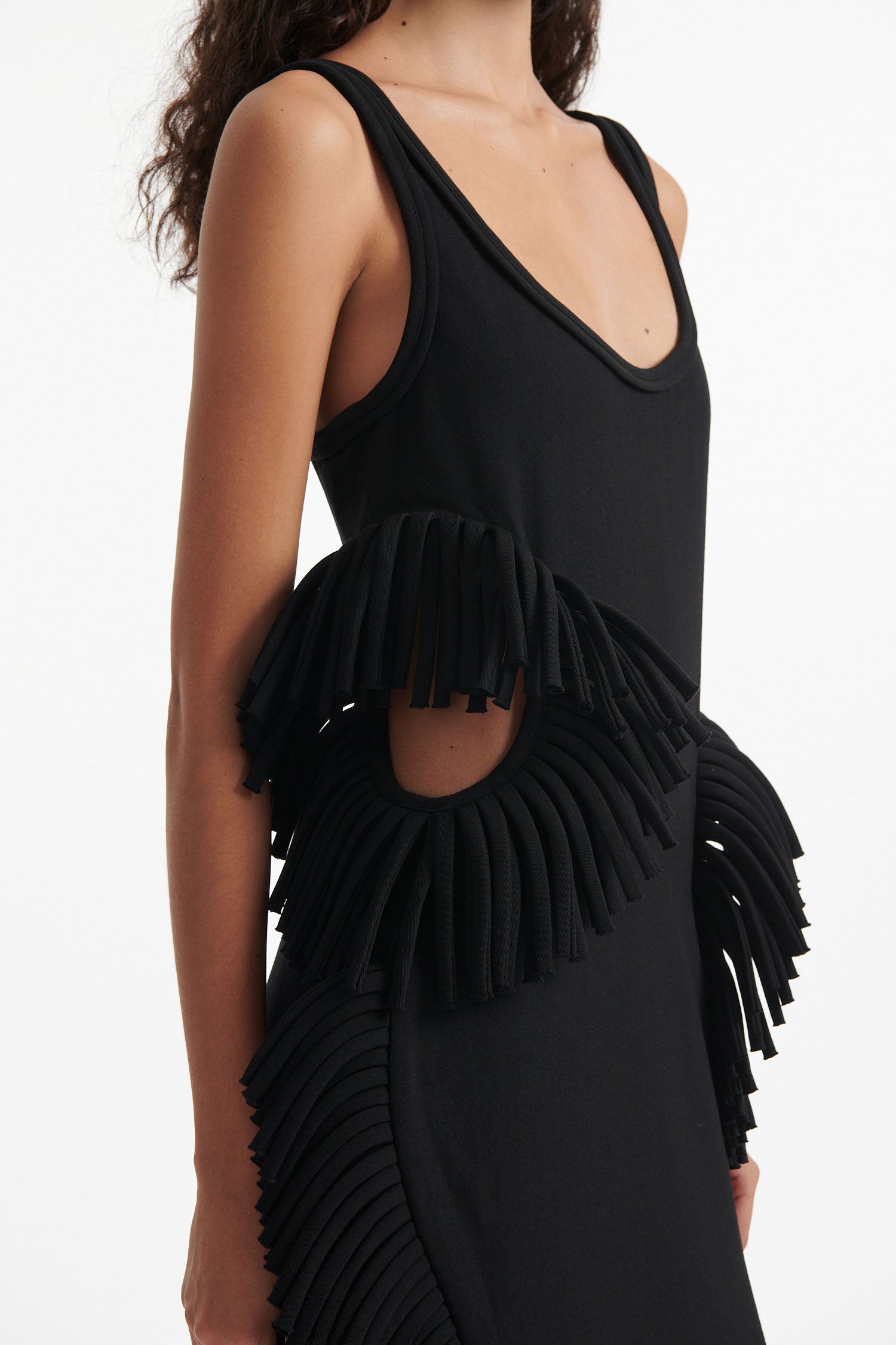 Female model wears black midi tank dress with large fringe openings at either side and scoop fringe hem with black high heel boots on white background.
