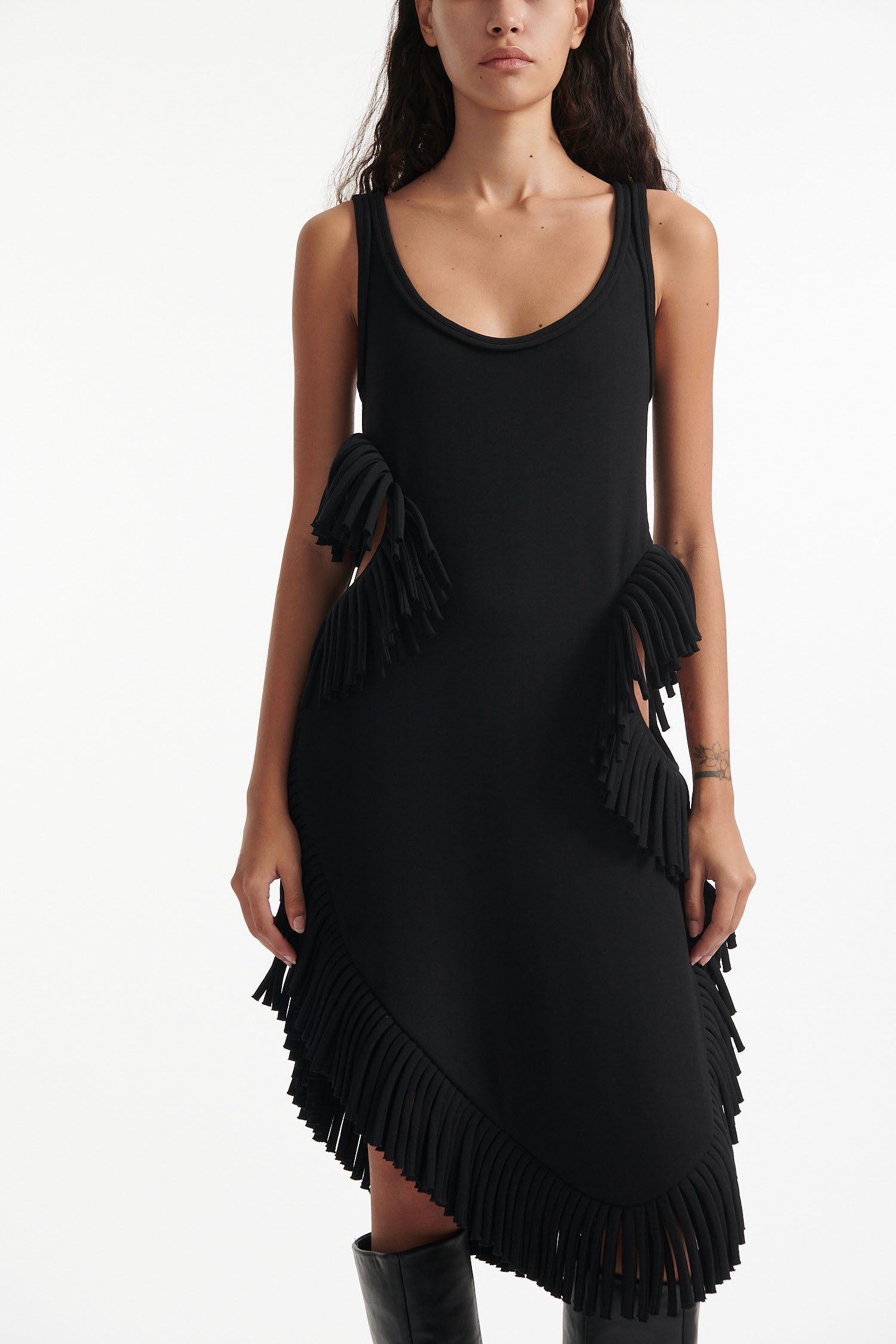 Female model wears black midi tank dress with large fringe openings at either side and scoop fringe hem with black high heel boots on white background.