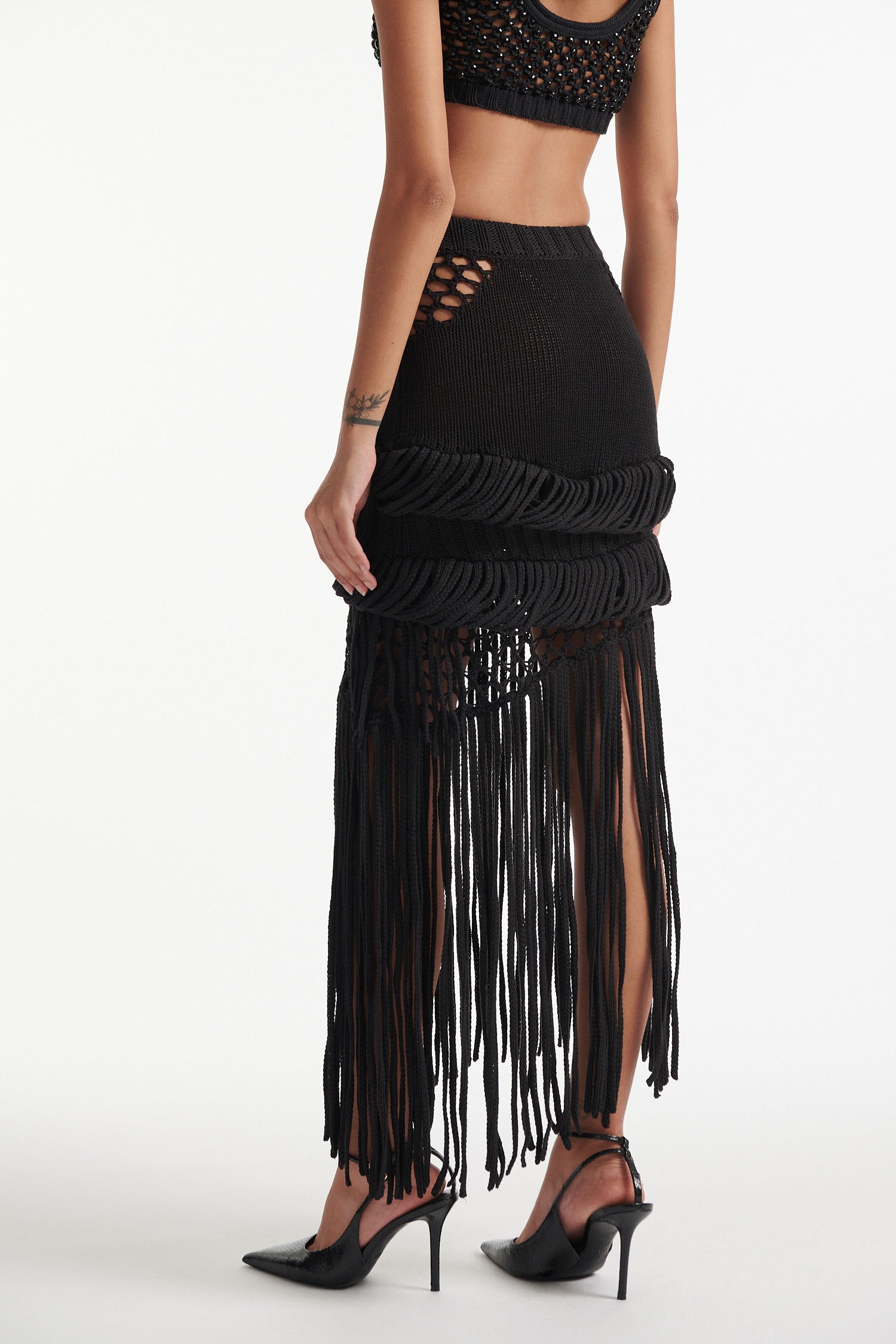 Female model wears long black fringe mixed-stitch see-through skirt with black crystal embellished bralette and black high heels on white background. 