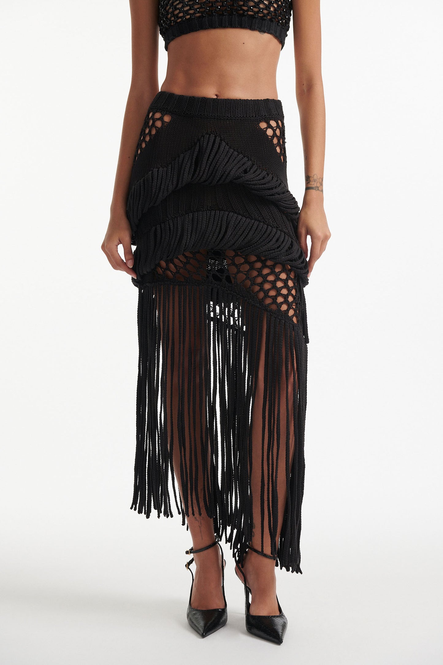 Female model wears long black fringe mixed-stitch see-through skirt with black crystal embellished bralette and black high heels on white background. 