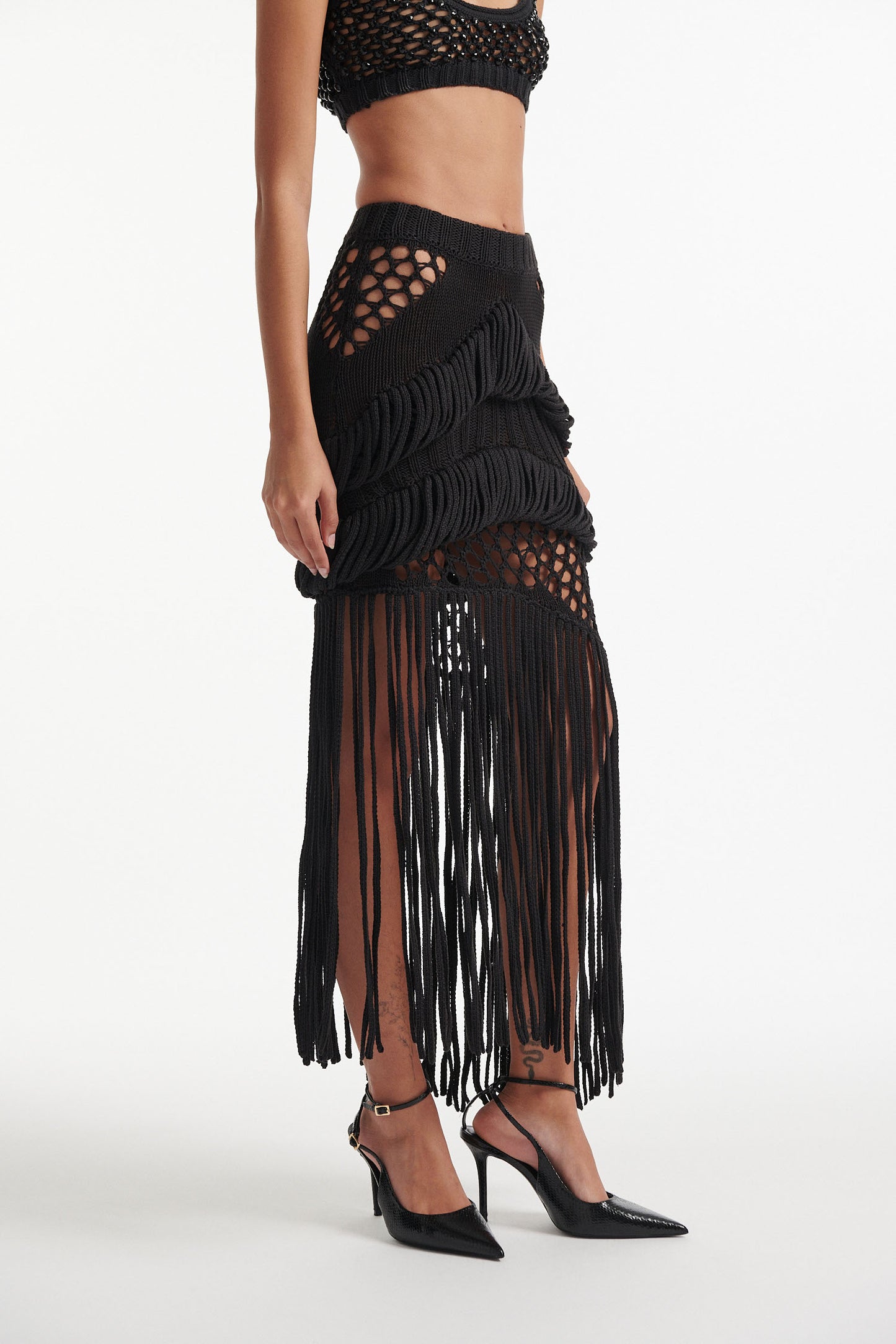 Female model wears long black fringe mixed-stitch see-through skirt with black crystal embellished bralette and black high heels on white background. 