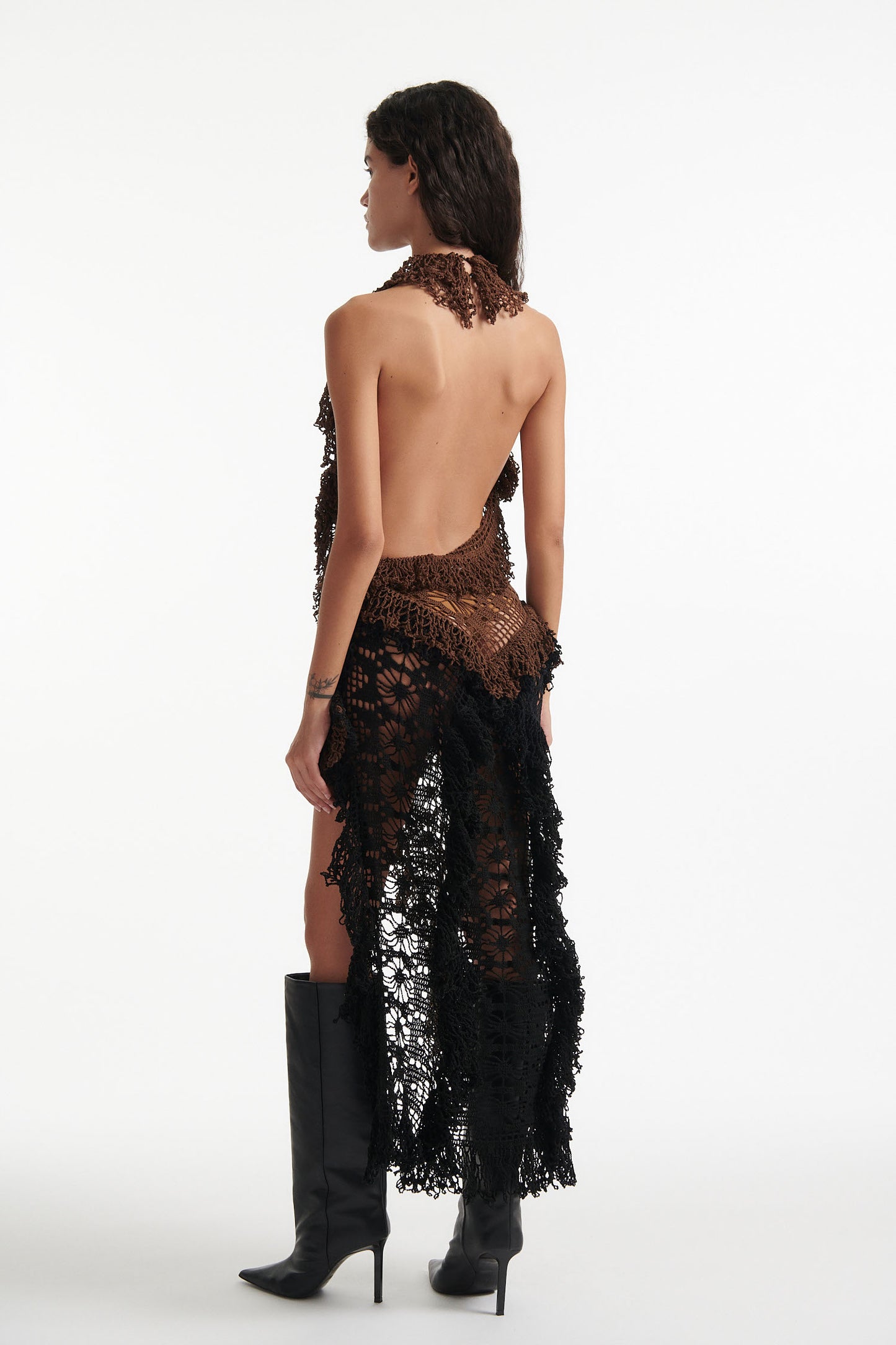 Female model wears open-back brown and black hand knit crochet dress with black high heels on white background. The front of the dress is brown and the back of the dress is black. 