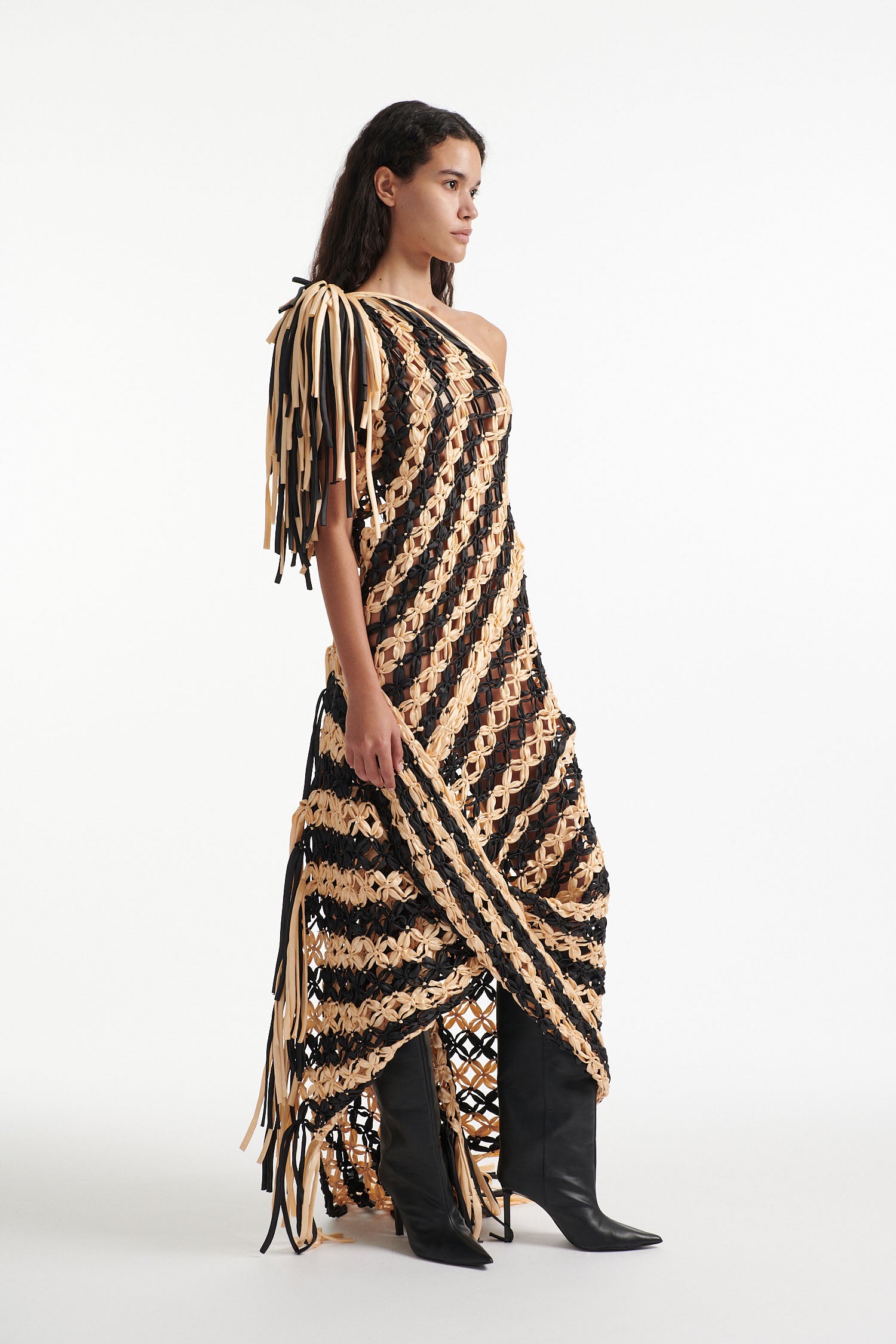 Female model wears off-the-shoulder black and cream knotted macrame gown with black high heel boots on white background. The macrame is stitched in a wide diagonal stripe and ends in fringe on the shoulder and hem. 