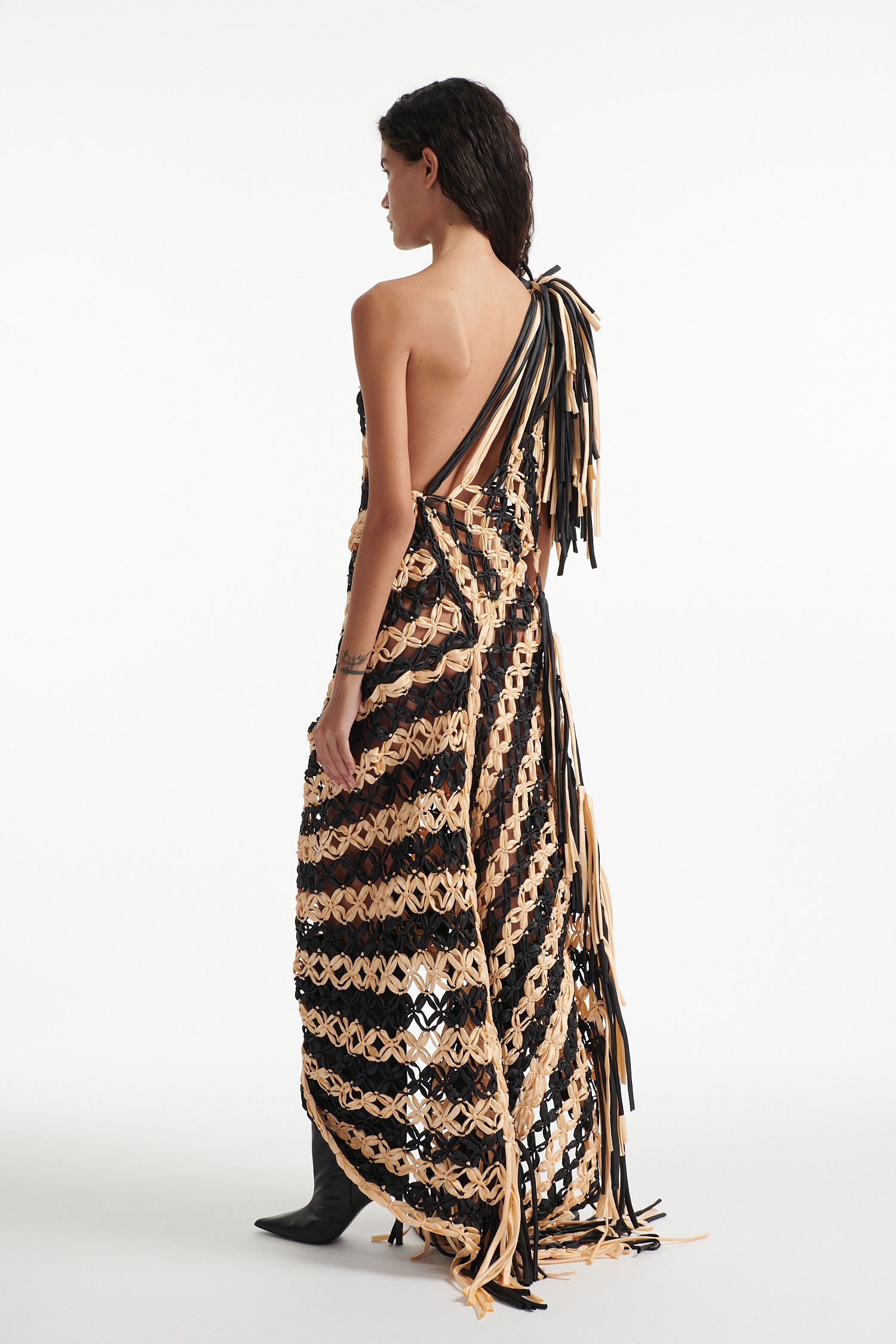 Female model wears off-the-shoulder black and cream knotted macrame gown with black high heel boots on white background. The macrame is stitched in a wide diagonal stripe and ends in fringe on the shoulder and hem. 