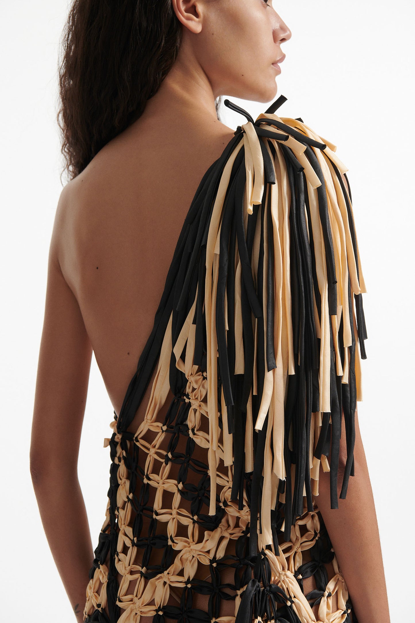 Female model wears off-the-shoulder black and cream knotted macrame gown with black high heel boots on white background. The macrame is stitched in a wide diagonal stripe and ends in fringe on the shoulder and hem. 