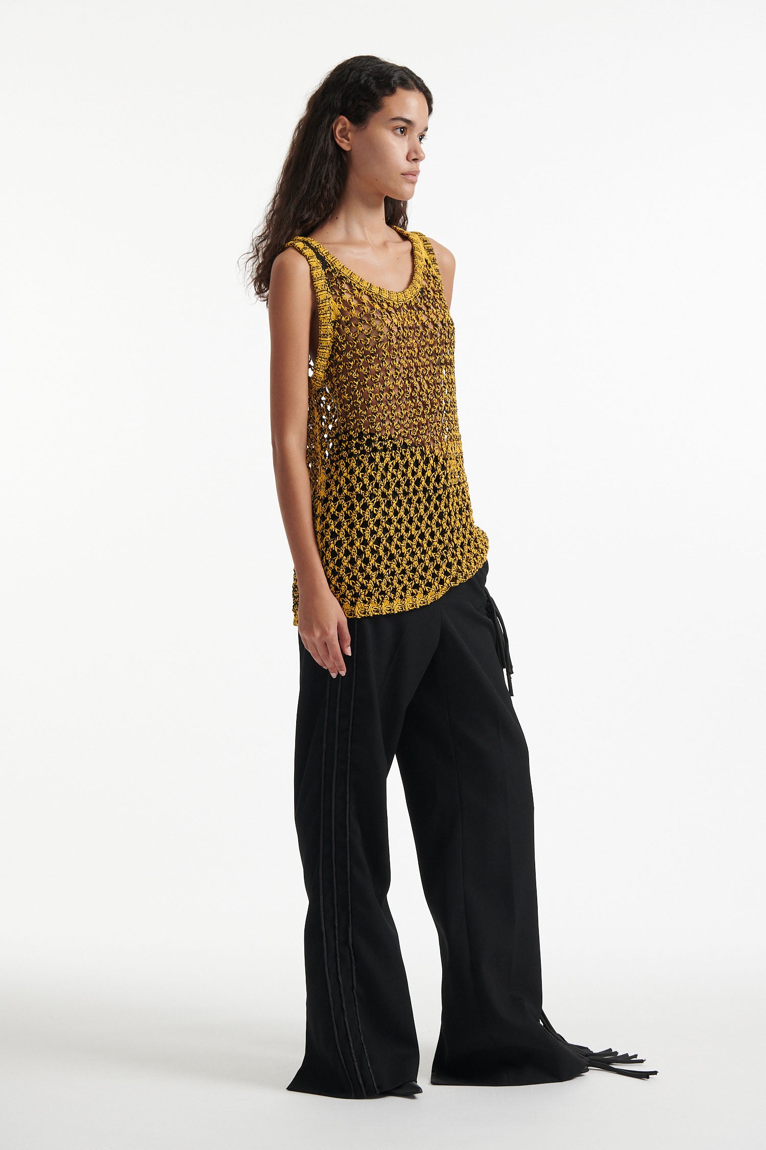 Female model wears yellow mesh tank dress, black fringe trousers, black high heel boots, and chunky silver bracelet on white background. 