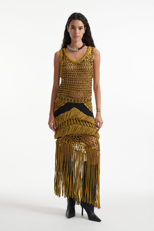 Female model wears yellow mesh tank and matching mixed-stitch skirt with black high heel boots on white background. 