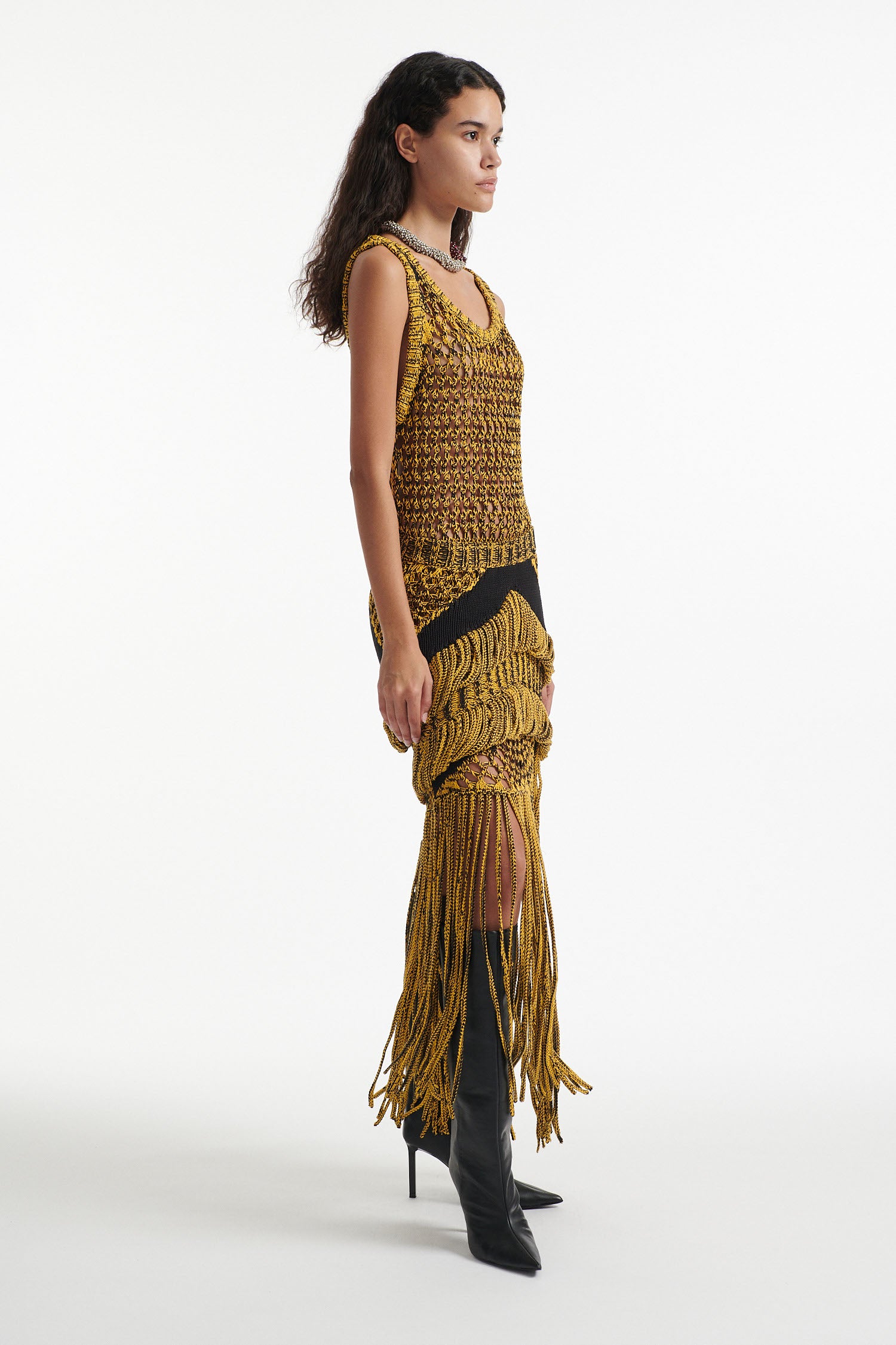 Female model wears yellow mesh tank and matching mixed-stitch skirt with black high heel boots on white background. 