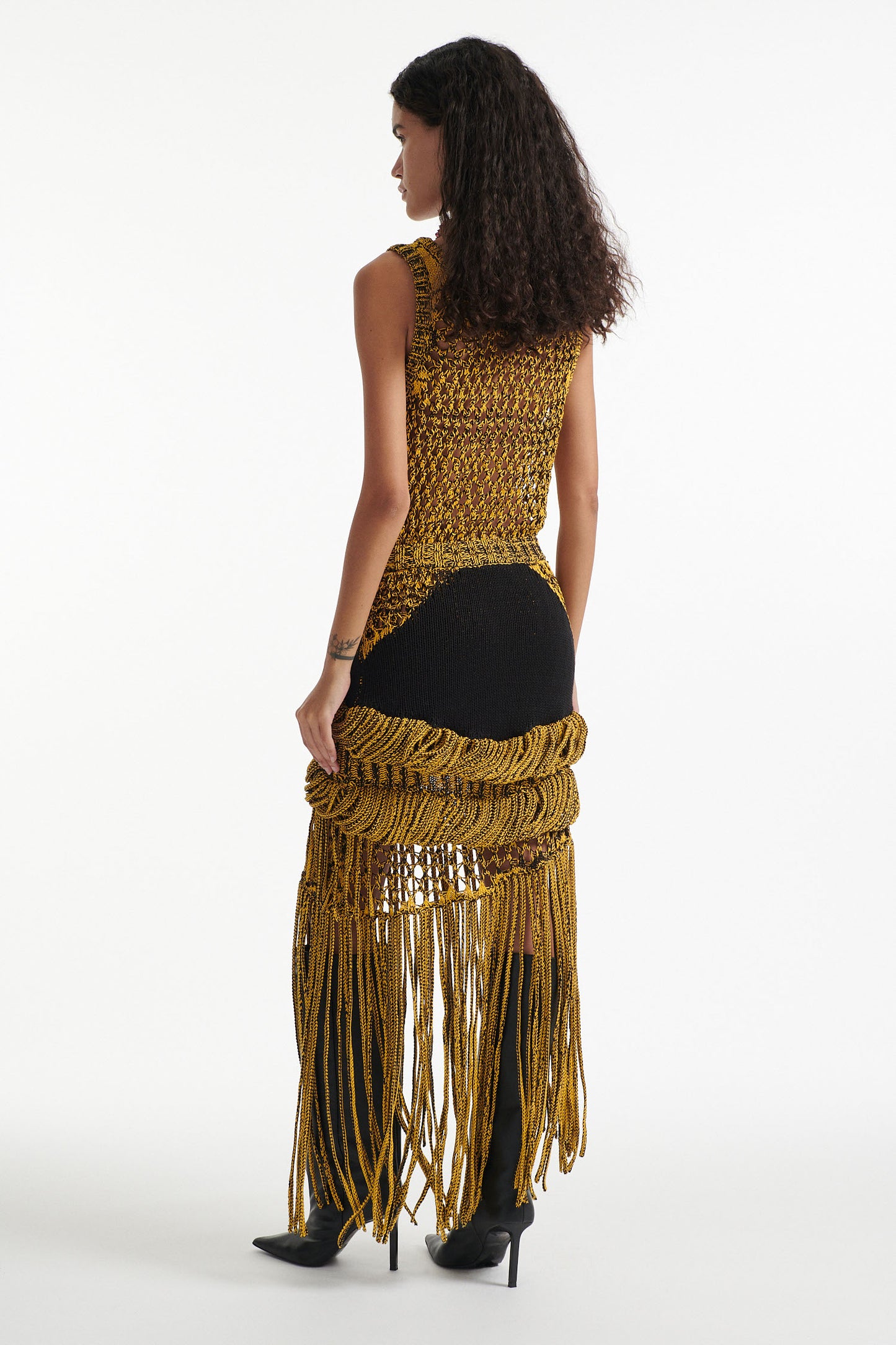 Female model wears yellow mesh tank and matching mixed-stitch skirt with black high heel boots on white background. 