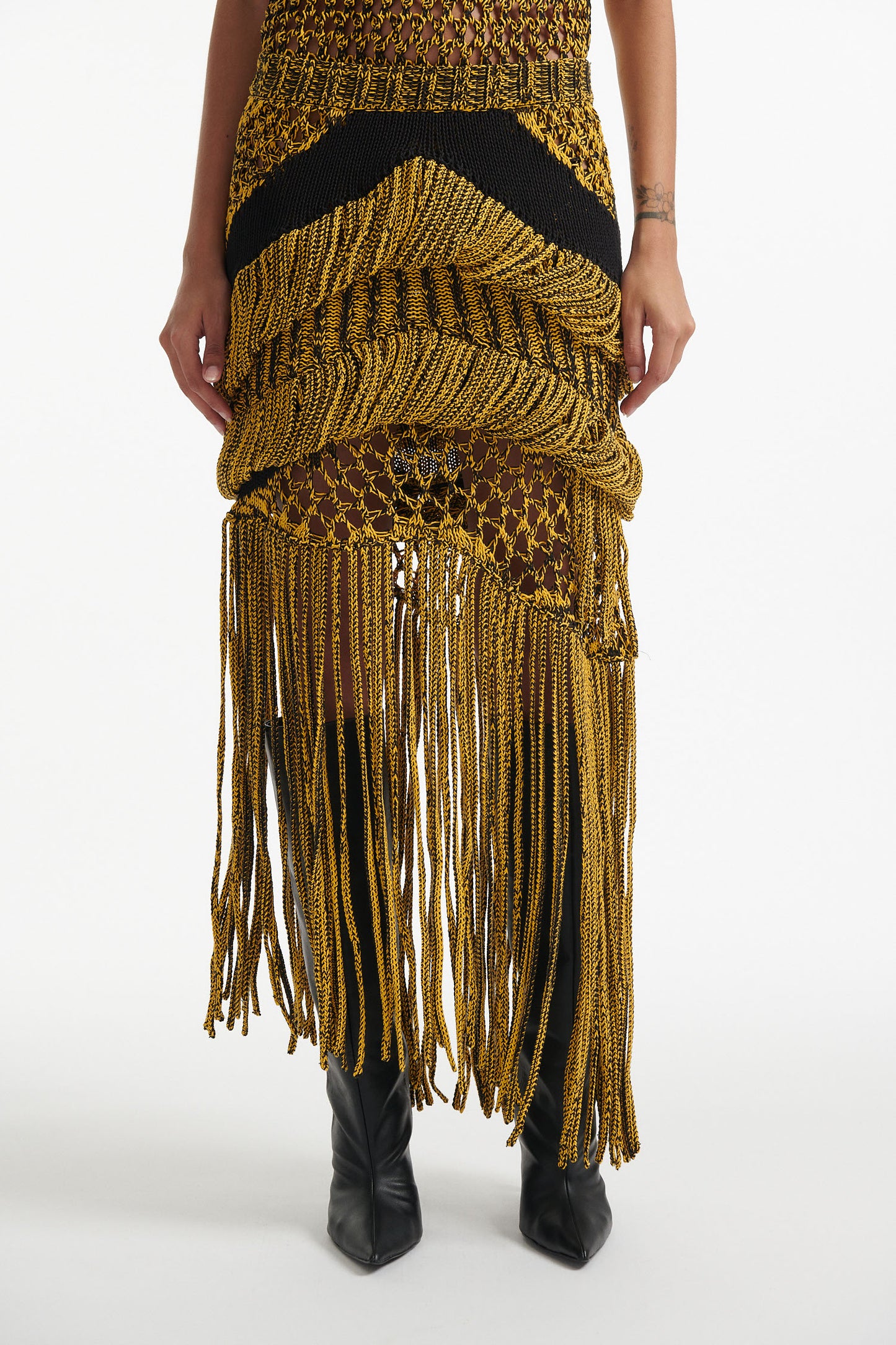 Female model wears yellow mesh tank and matching mixed-stitch skirt with black high heel boots on white background. 