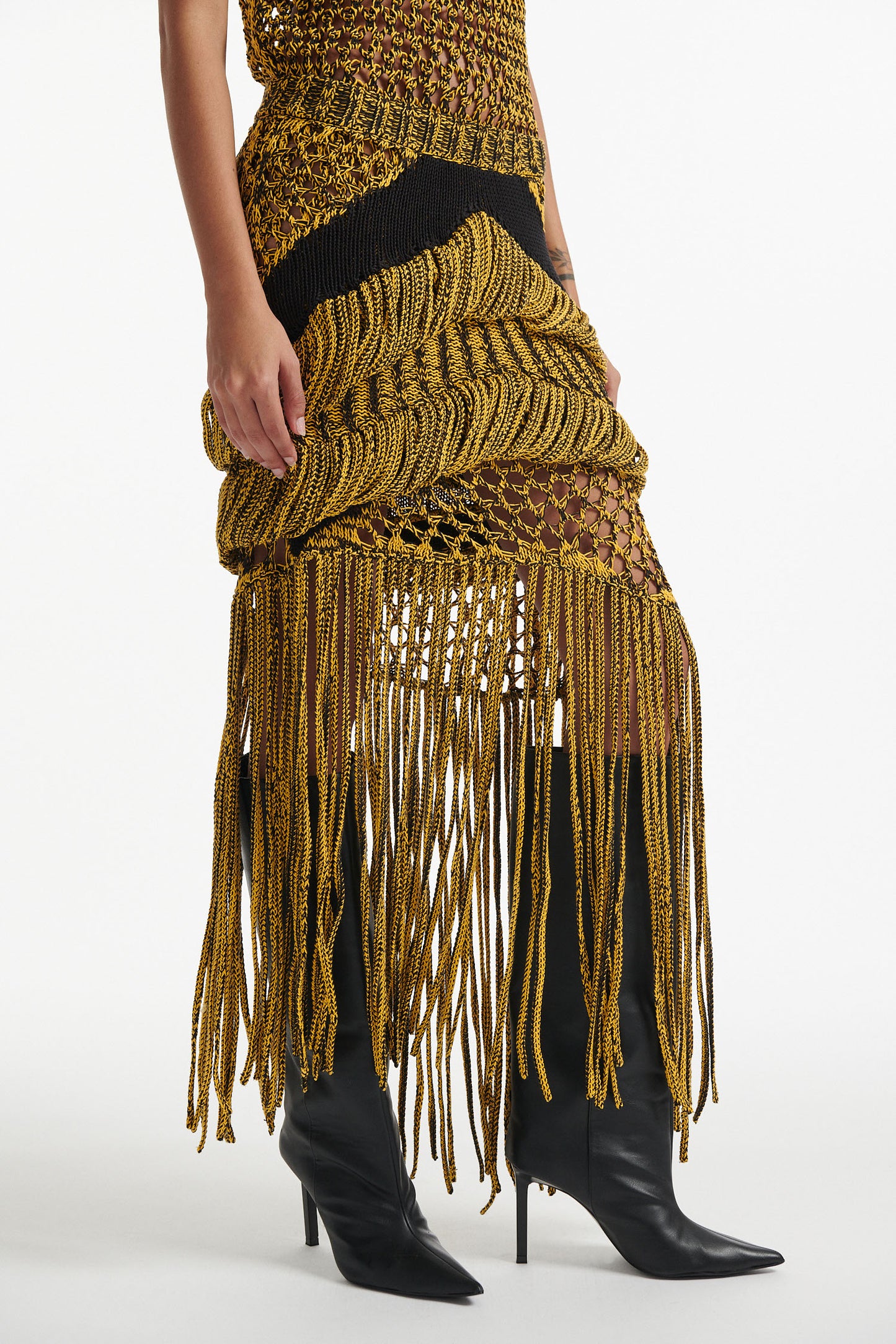 Female model wears yellow mesh tank and matching mixed-stitch skirt with black high heel boots on white background. 