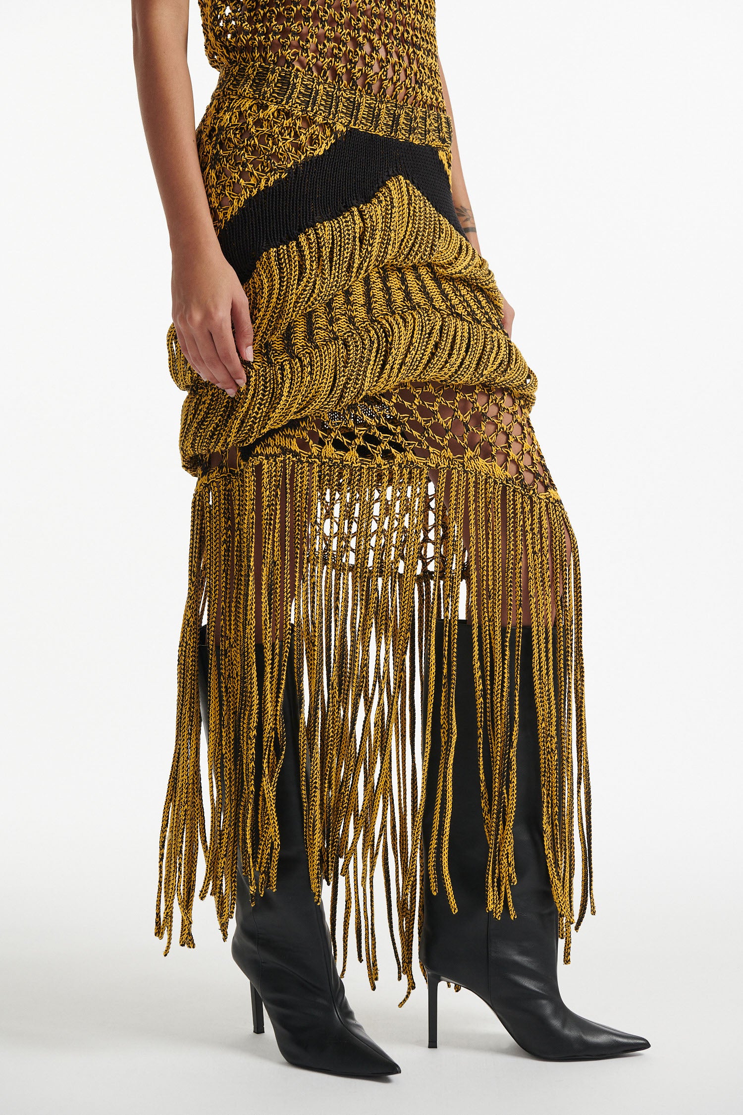 Female model wears yellow mesh tank and matching mixed-stitch skirt with black high heel boots on white background. 