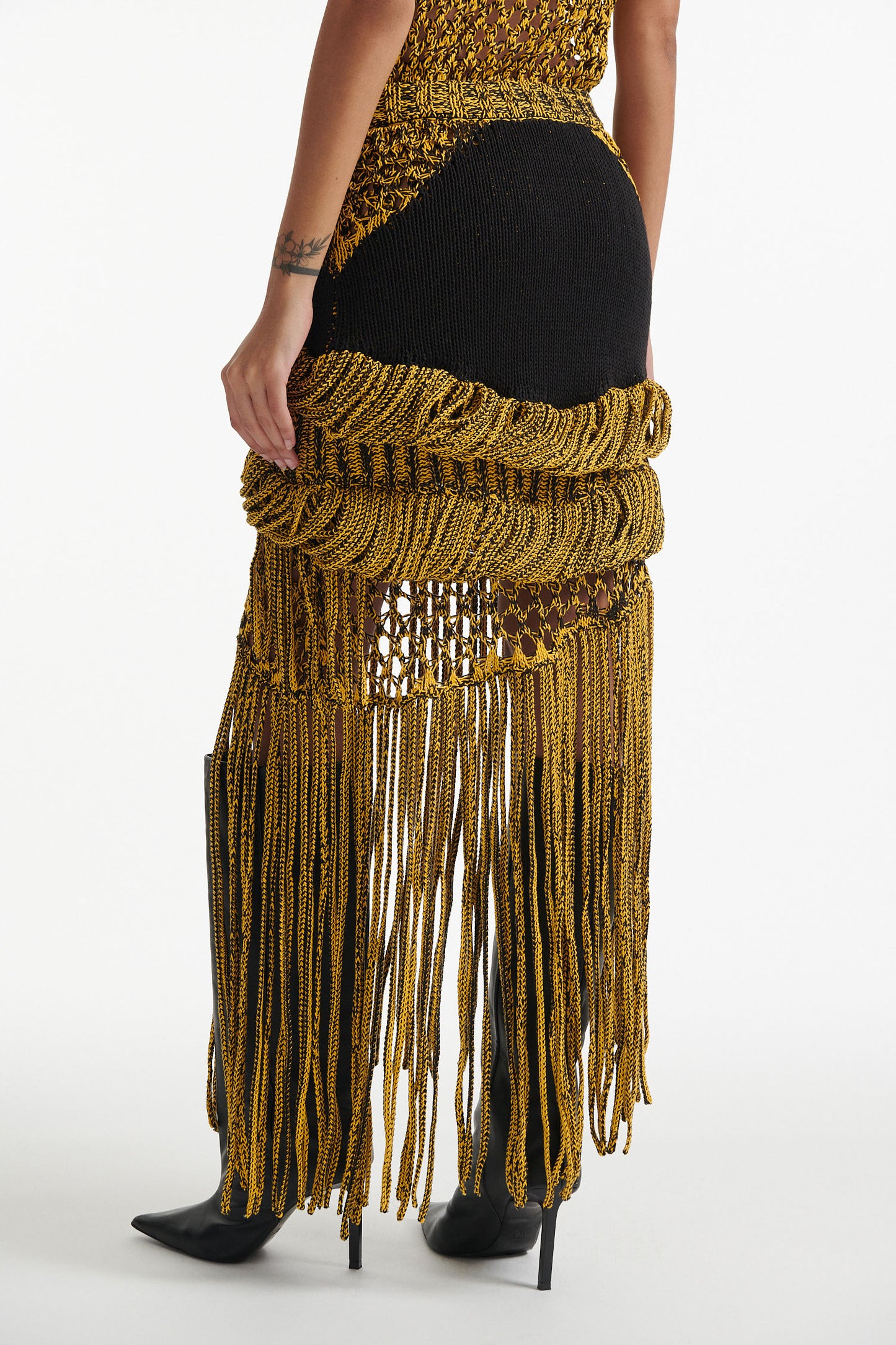 Female model wears yellow mesh tank and matching mixed-stitch skirt with black high heel boots on white background. 